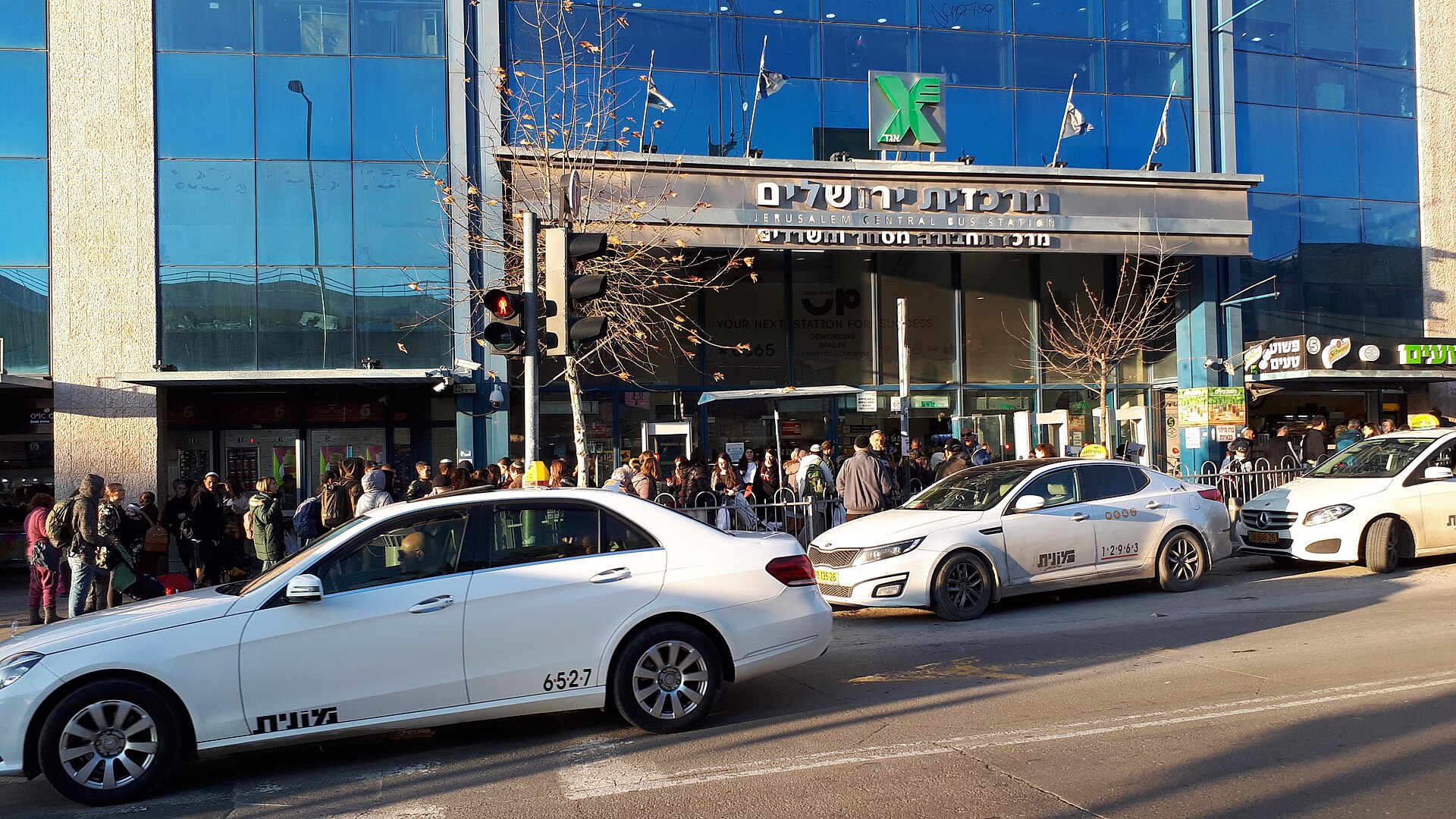Three white taxis with yellow roof signs and Hebrew taxi lettering wait outside the glass-fronted Jerusalem Central Bus Station on Jaffa Road