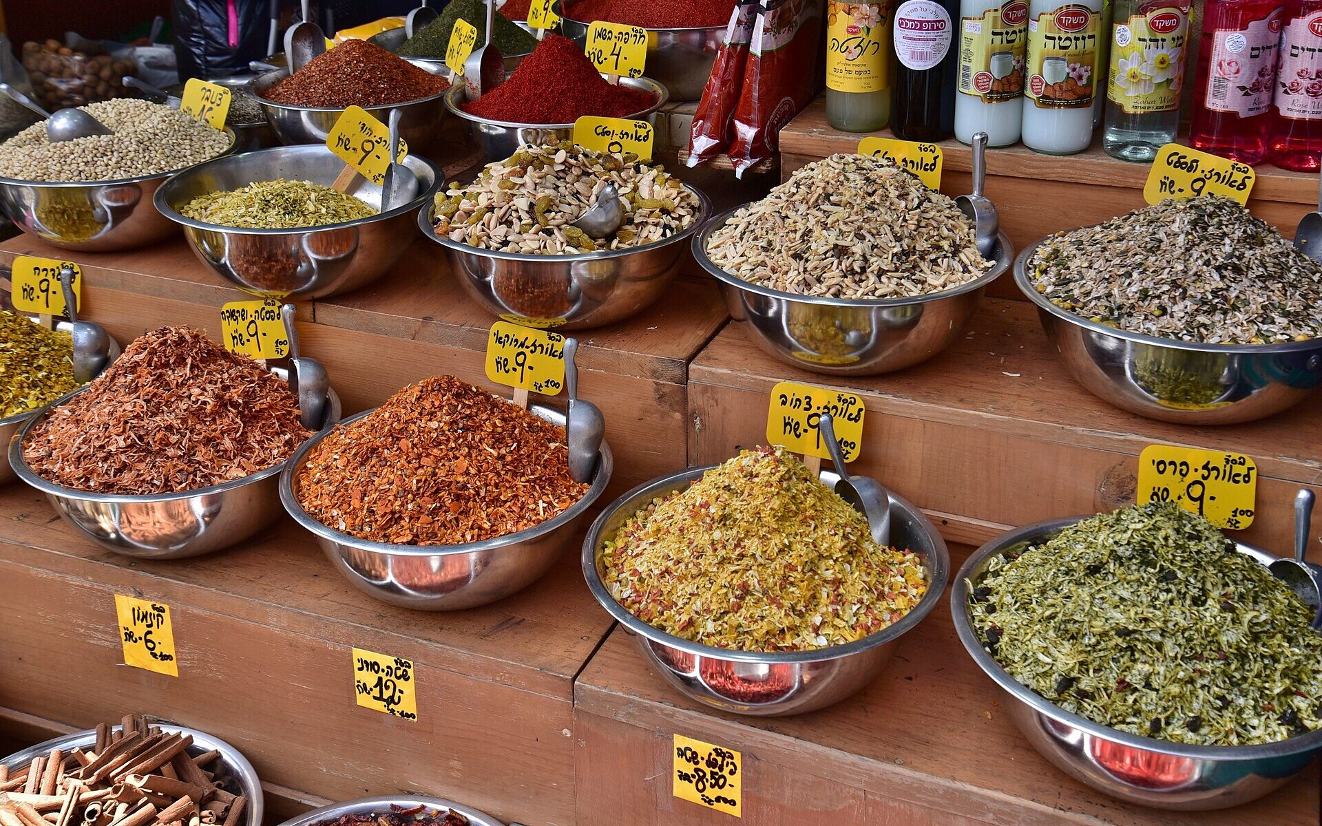 Tiered steel bowls of spices and rice seasoning blends with handwritten Hebrew price tags at a Mahane Yehuda spice stall, beside bottles of rose and orange blossom water