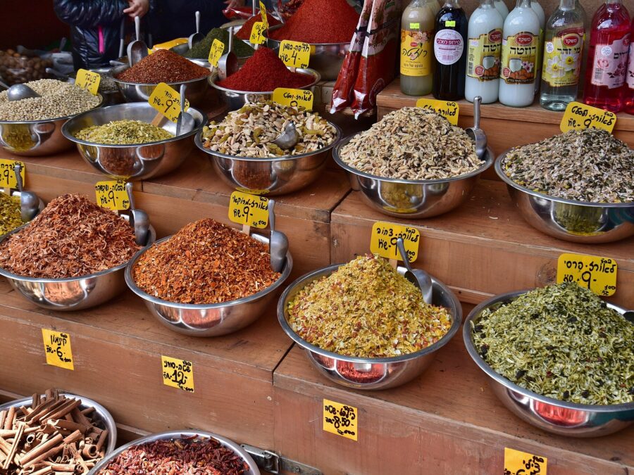 Tiered steel bowls of spices and rice seasoning blends with handwritten Hebrew price tags at a Mahane Yehuda spice stall, beside bottles of rose and orange blossom water