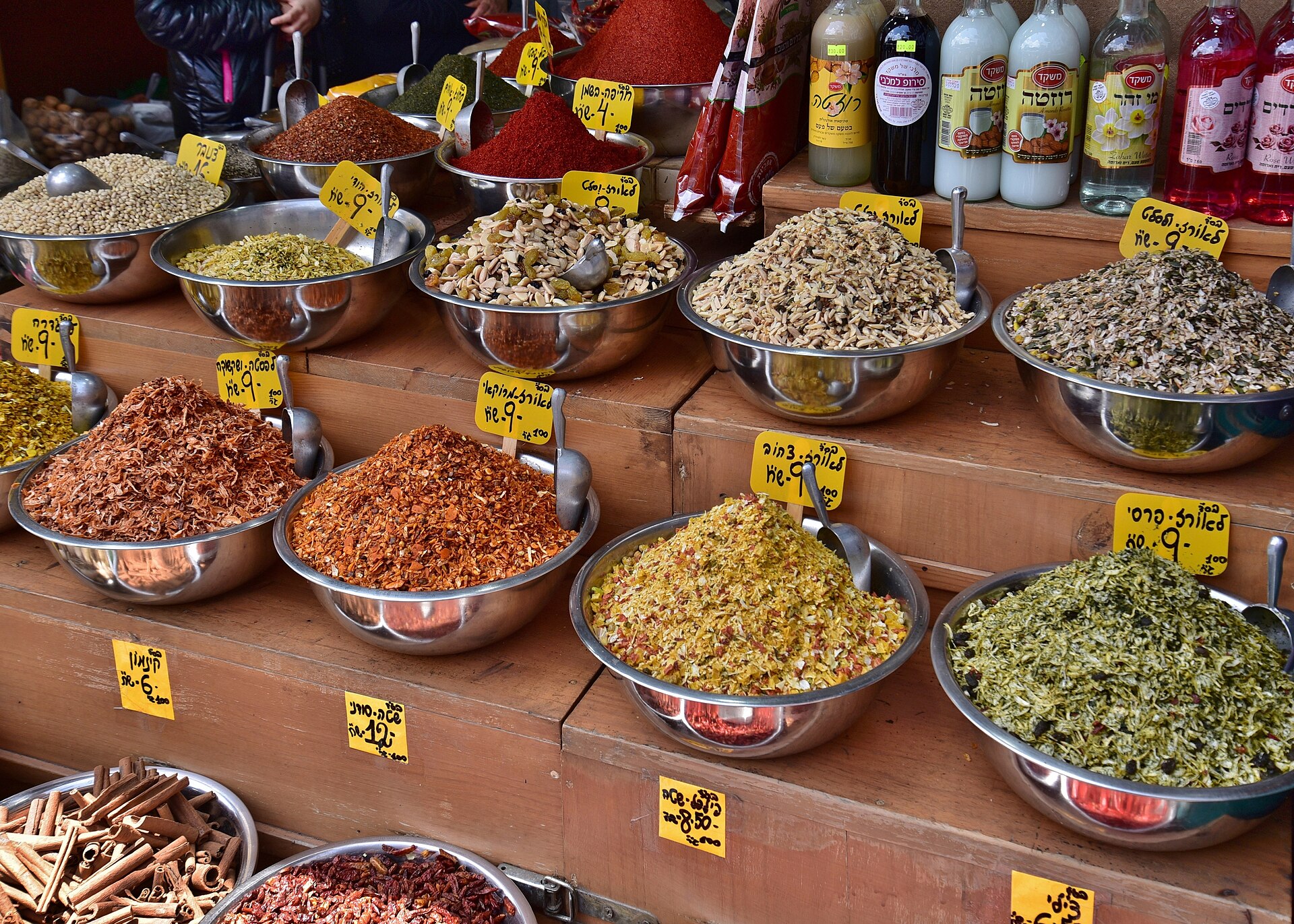 Tiered steel bowls of spices and rice seasoning blends with handwritten Hebrew price tags at a Mahane Yehuda spice stall, beside bottles of rose and orange blossom water