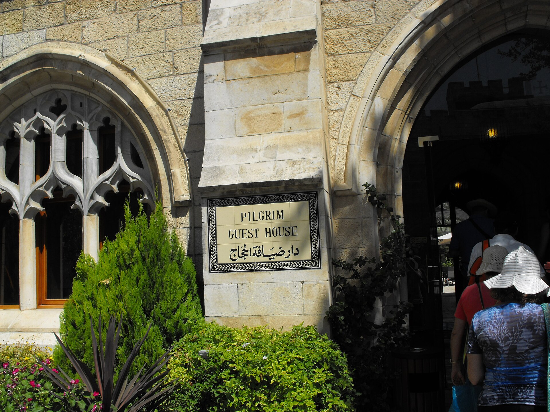 Tiled Pilgrim Guest House sign in English and Arabic on the stone wall of St George's Cathedral guesthouse in Jerusalem, between a Gothic traceried window and an arched gateway