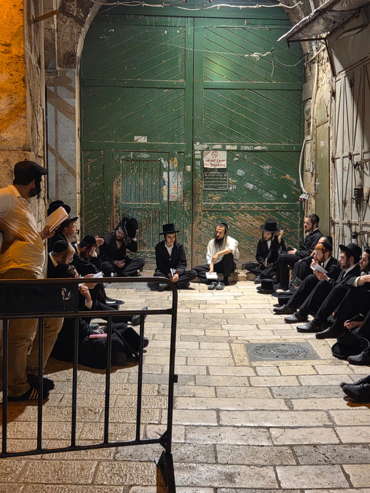 Ultra-Orthodox men in black hats sitting on the stone paving of an Old City alley at night, reading from small booklets in front of a large green gate