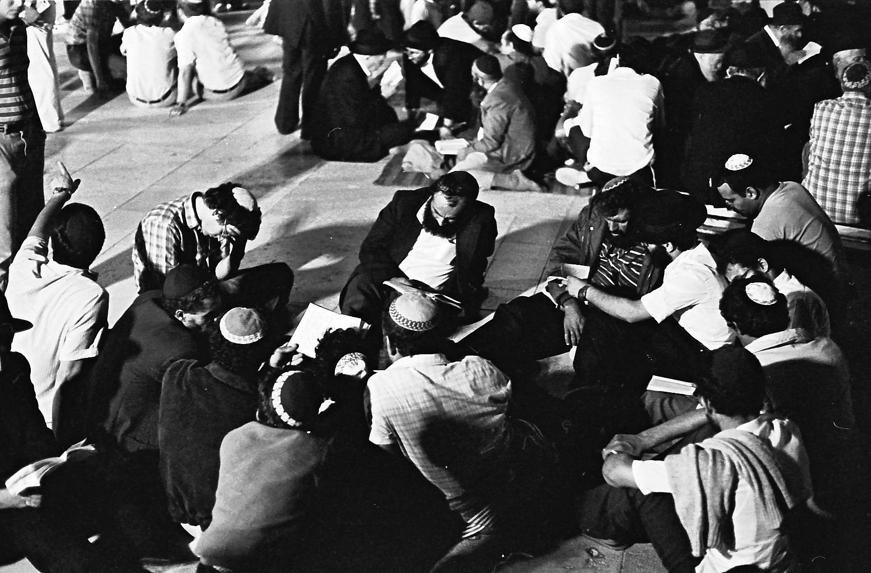 Black and white photograph of men in skullcaps sitting in a circle on the ground at the Western Wall plaza at night, reading from booklets