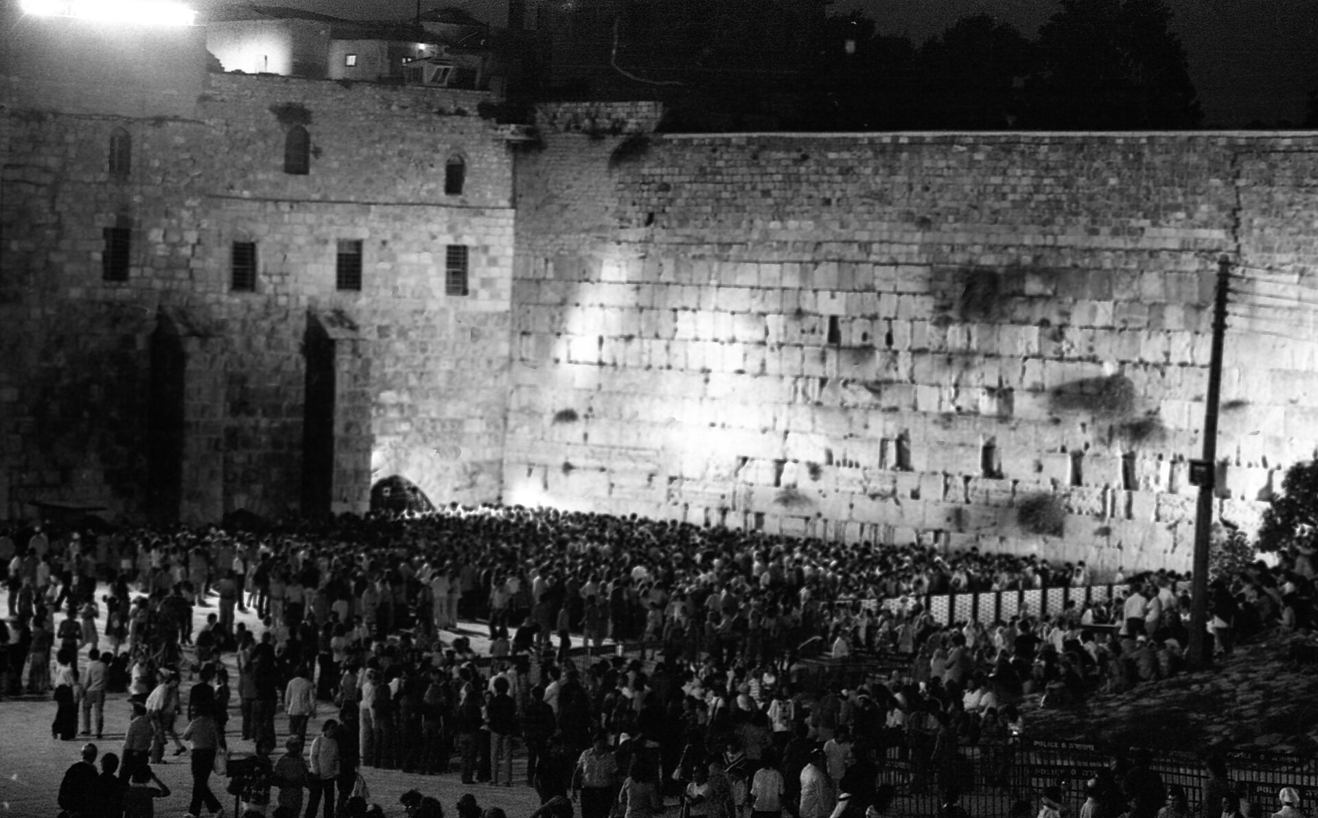 Black and white night photograph of a dense crowd filling the floodlit Western Wall plaza, with the great Herodian stones rising behind