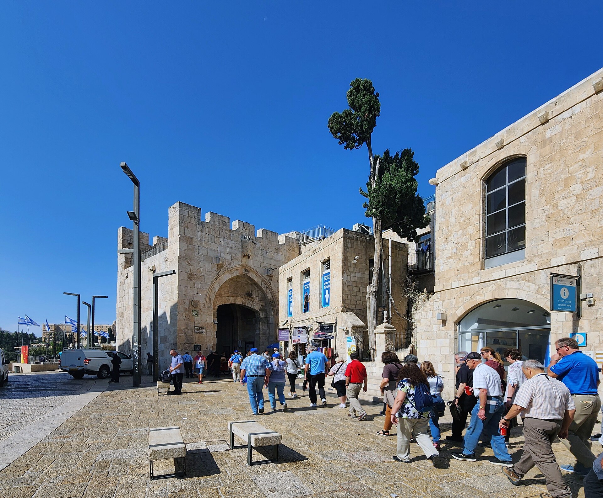 A tour group crossing the paved square just inside Jaffa Gate, passing the tourist information office and heading towards the gate's inner arch under a blue sky