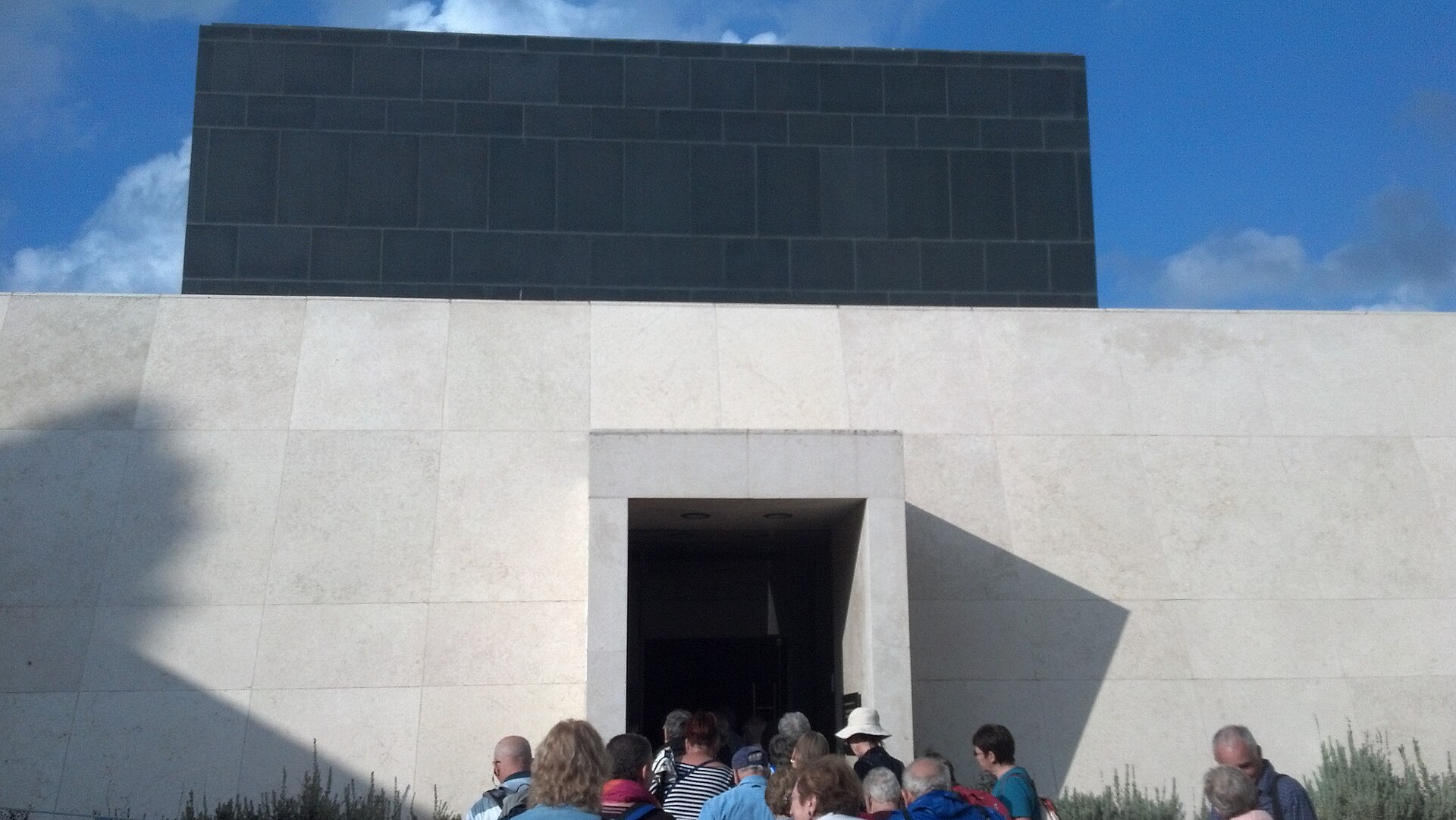 Tour group filing through a doorway in a white limestone wall into the Shrine of the Book, with the museum's black basalt wall rising above