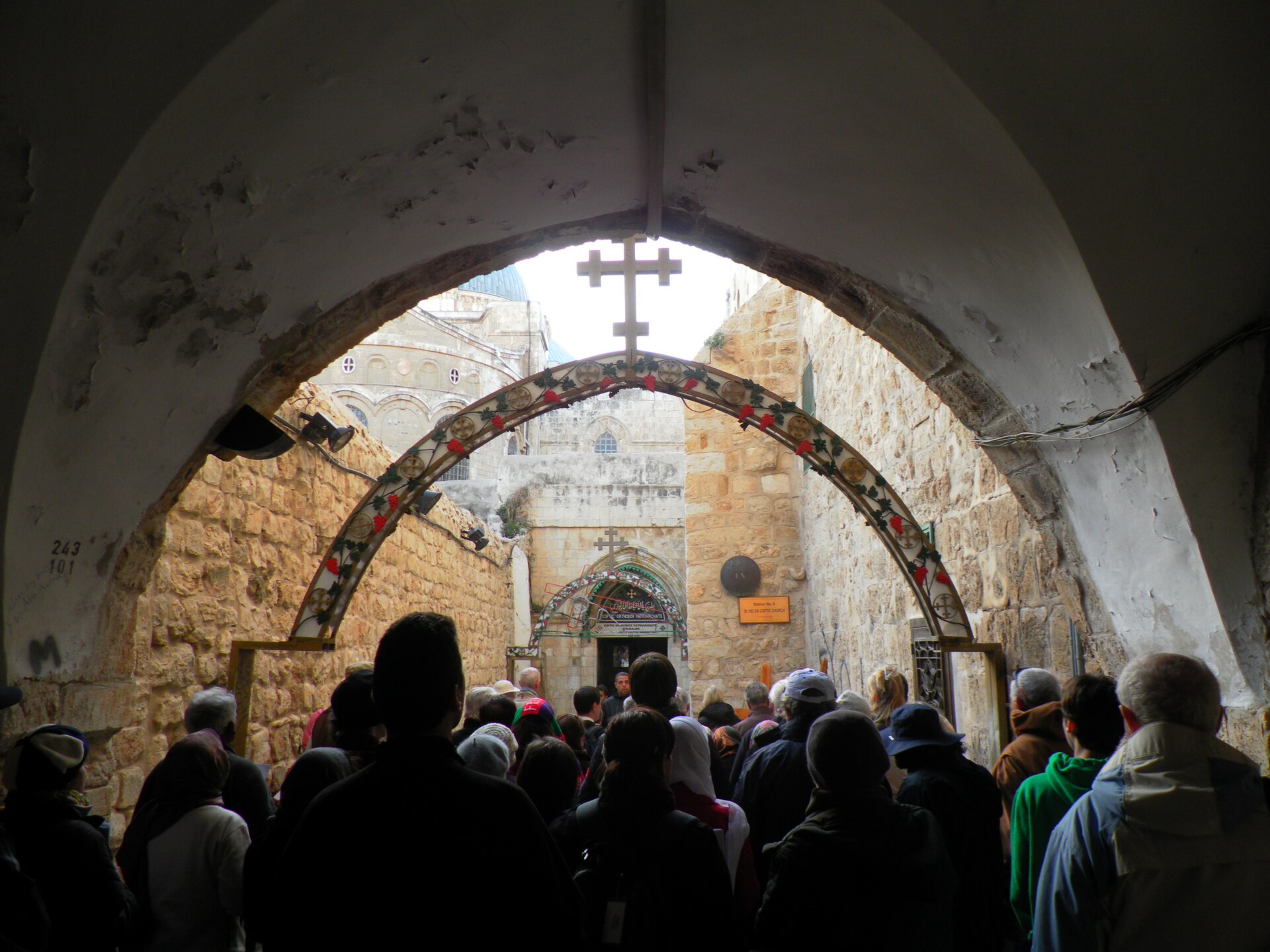A tour group of mostly older visitors, seen from behind, passes under a stone arch towards the Ninth Station of the Via Dolorosa below the Holy Sepulchre domes