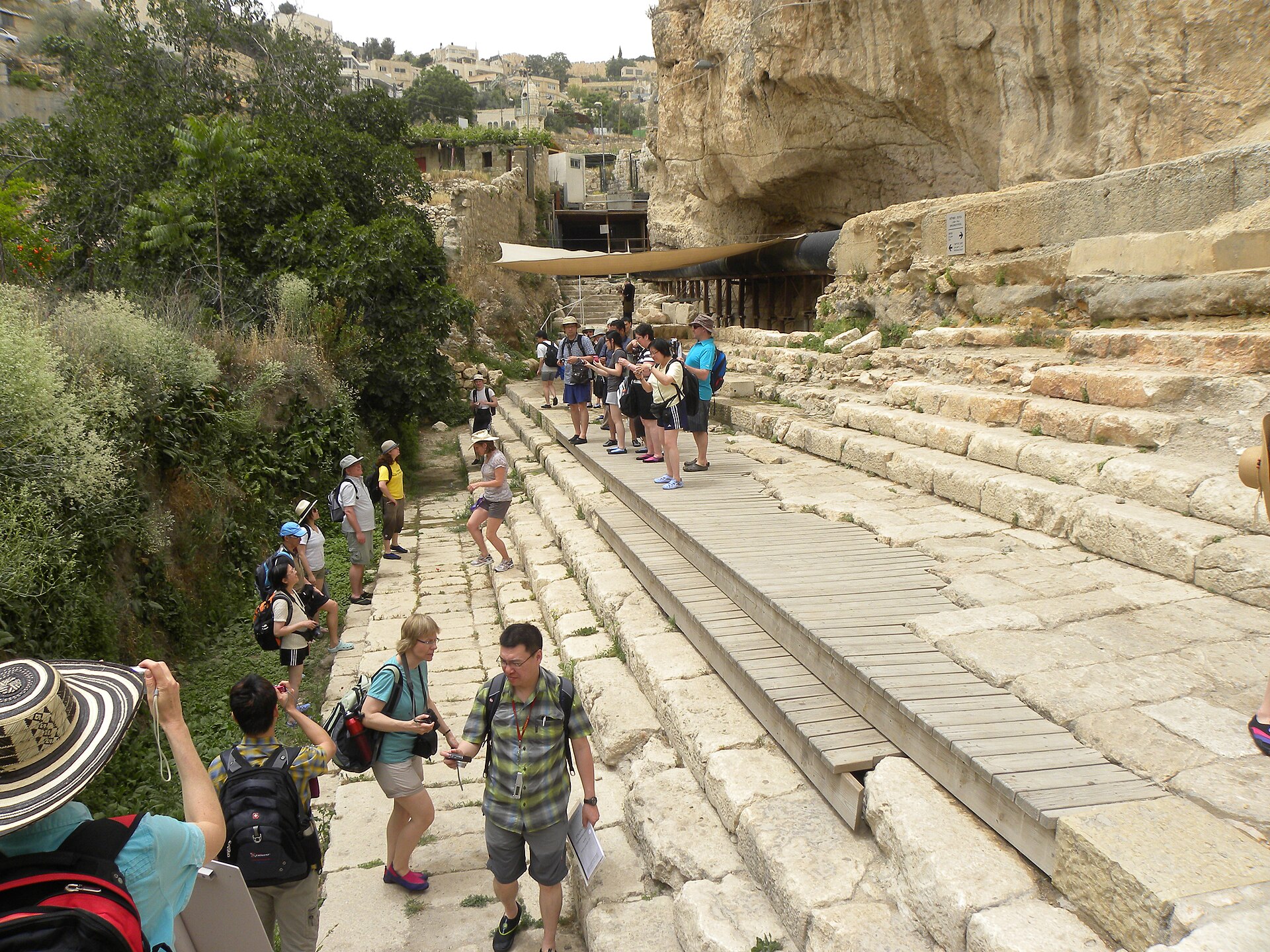 Tour group in sun hats and water shoes on the excavated stone steps and wooden boardwalk of the Pool of Siloam, below a rock face