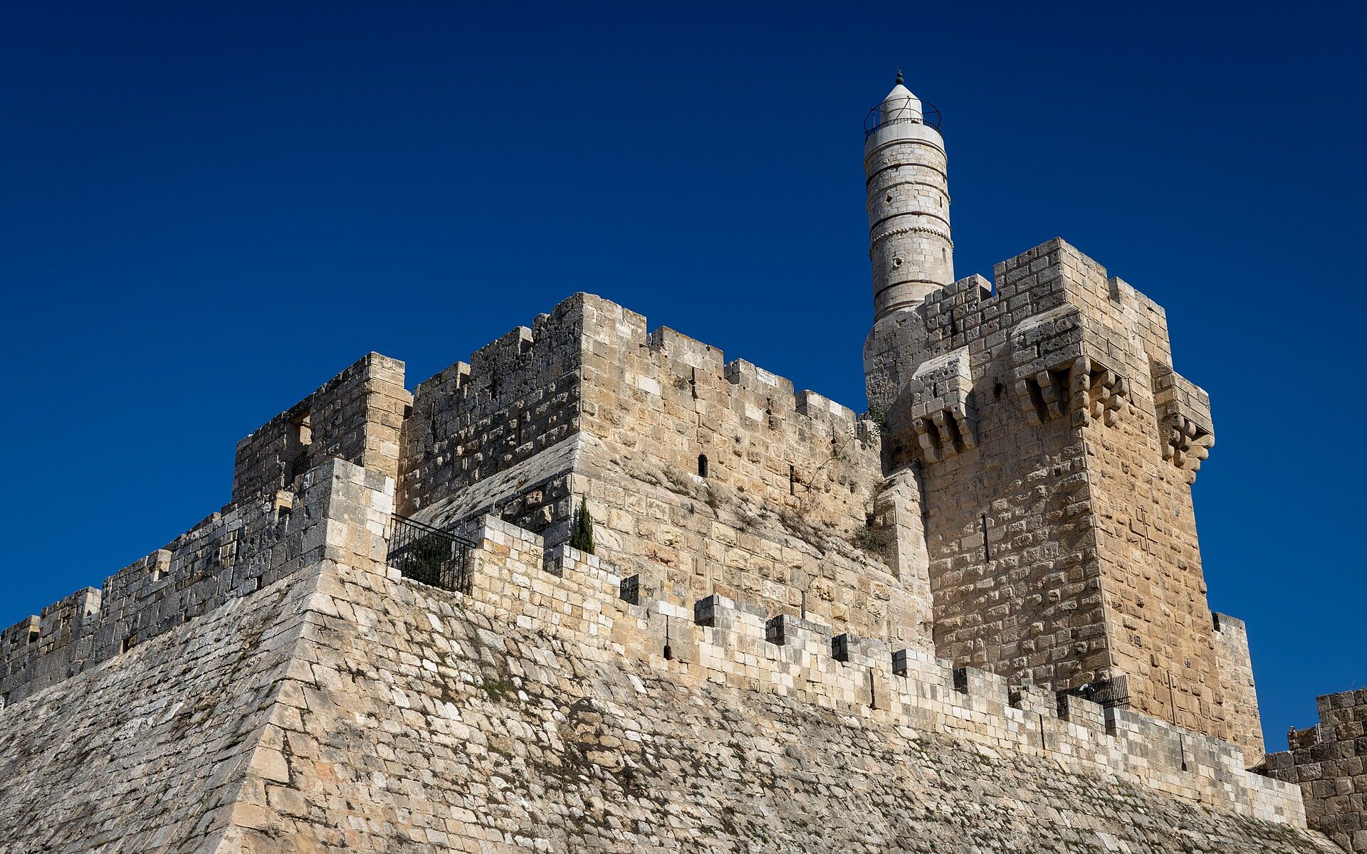 The Tower of David citadel in Jerusalem seen from below, its sloping stone glacis, crenellated ramparts and slender Ottoman minaret rising against a deep blue sky
