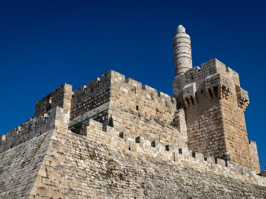 The Tower of David citadel in Jerusalem seen from below, its sloping stone glacis, crenellated ramparts and slender Ottoman minaret rising against a deep blue sky