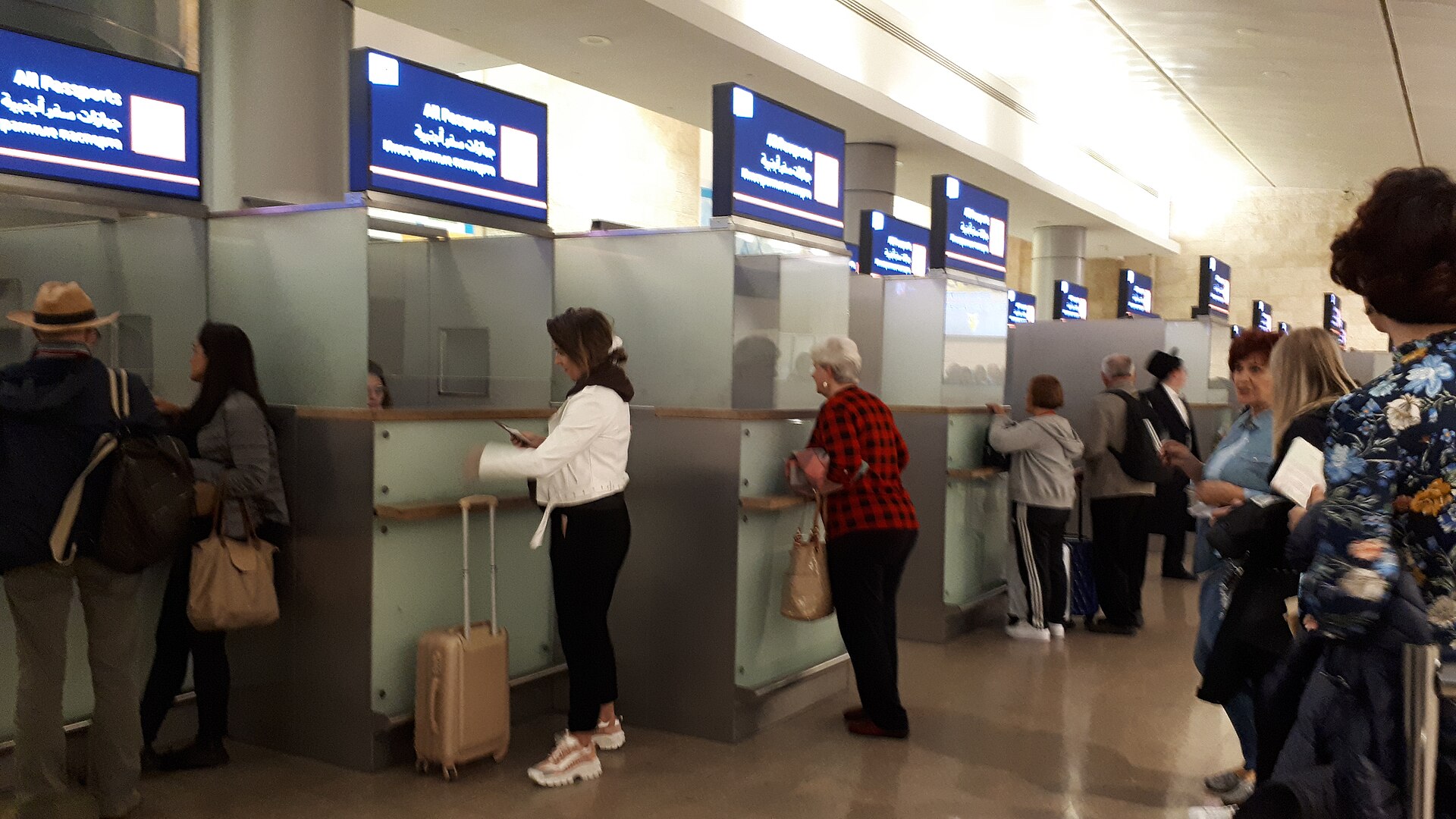 Travellers with passports and luggage stand at staffed passport control booths under blue All Passports signs in Ben Gurion Airport's Terminal 3