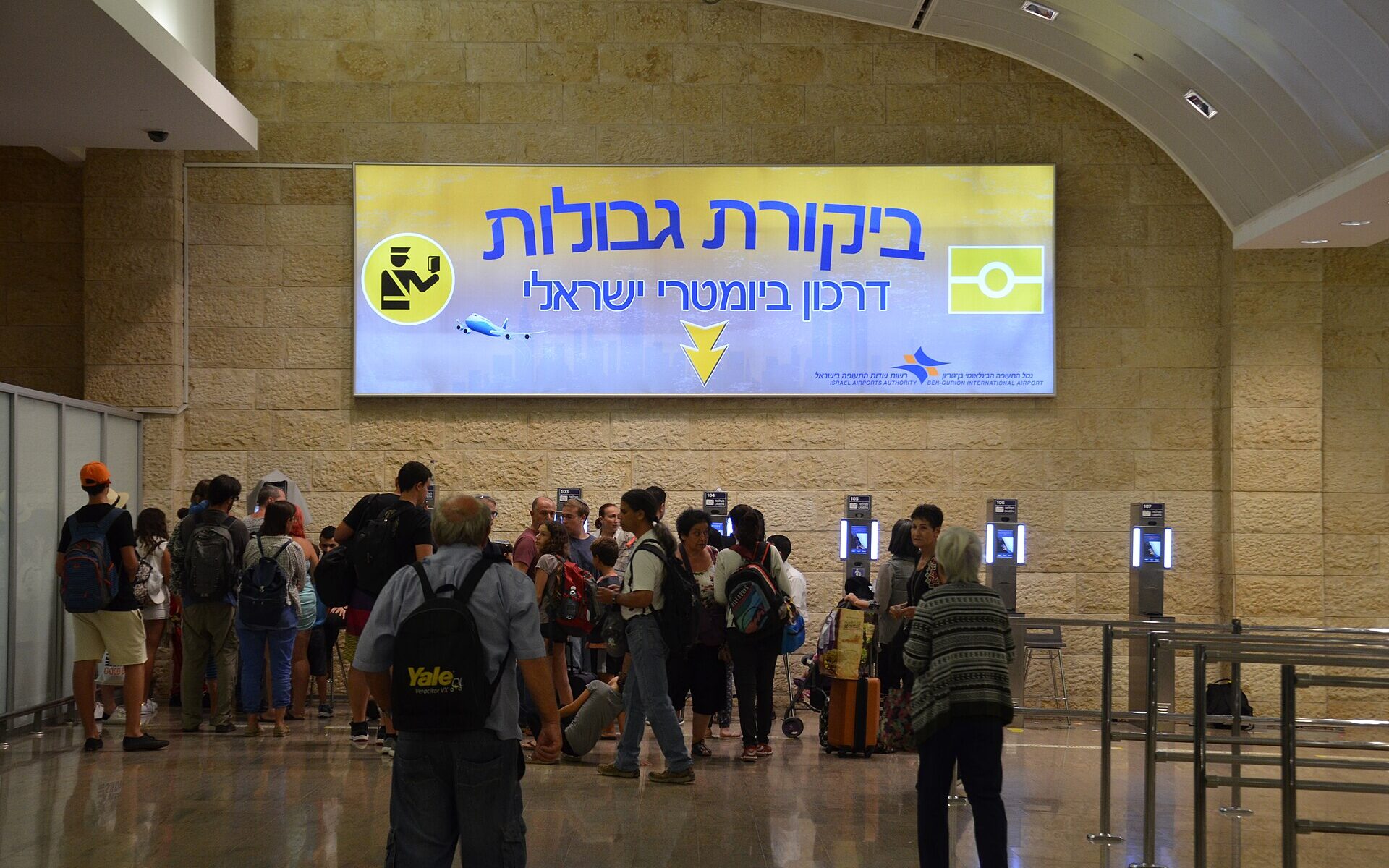 Travellers queue beneath a large Hebrew border control sign and a row of automated passport kiosks set into a Jerusalem-stone wall at Ben Gurion Airport
