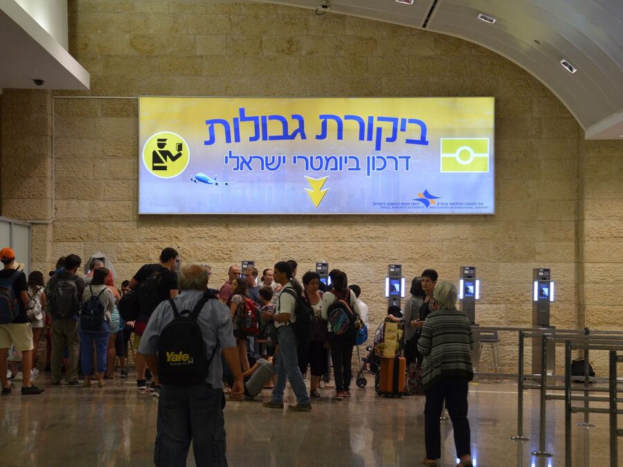 Travellers queue beneath a large Hebrew border control sign and a row of automated passport kiosks set into a Jerusalem-stone wall at Ben Gurion Airport