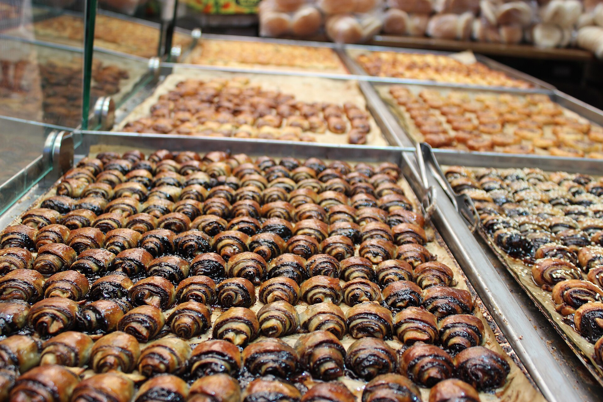 Trays of small, glossy chocolate rugelach lined up on a bakery counter in Mahane Yehuda Market, with more trays of rugelach and pastries behind