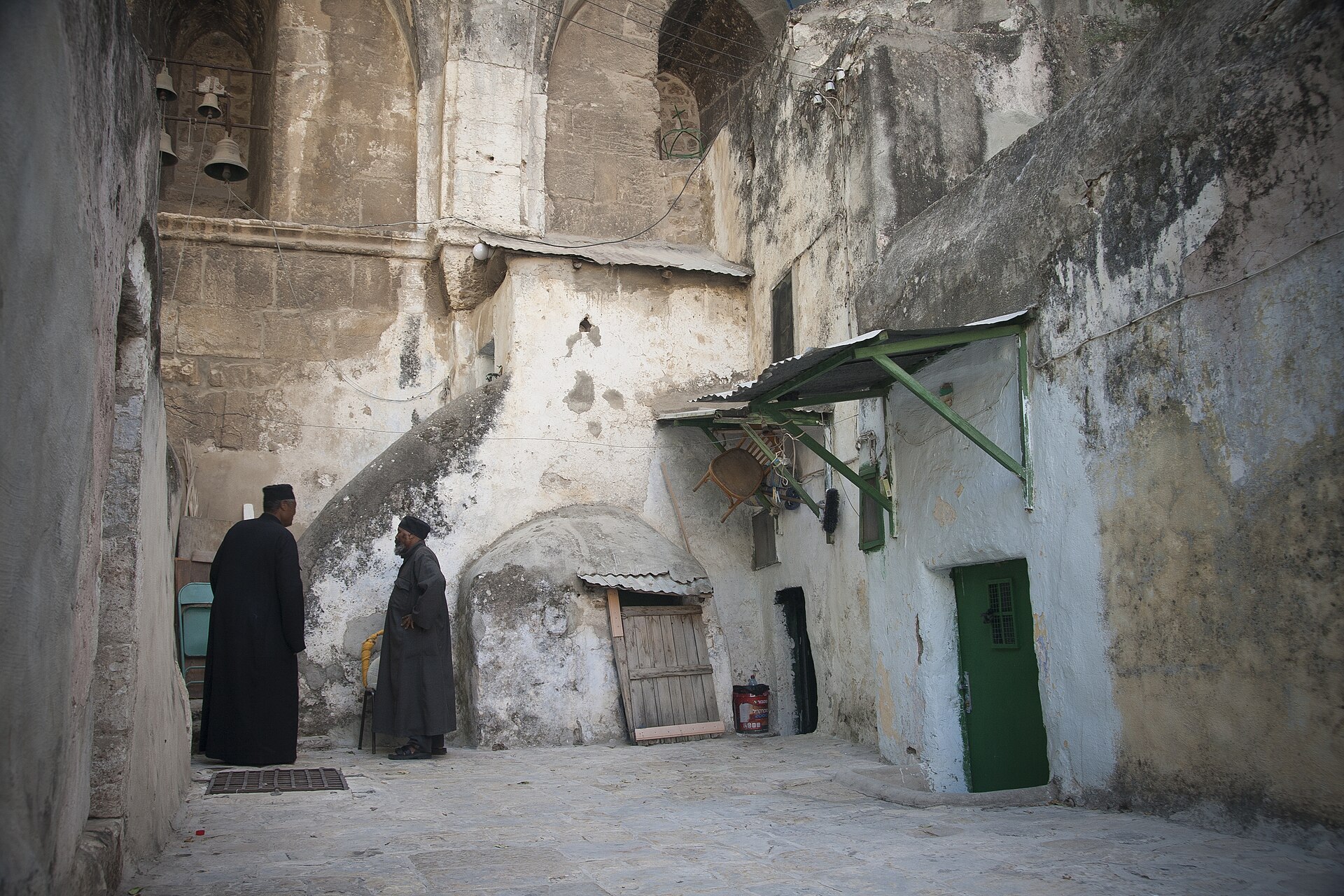 Two Ethiopian monks talking in a whitewashed courtyard of Deir es-Sultan, with small domed cells, green doors and bells hanging in the arches above
