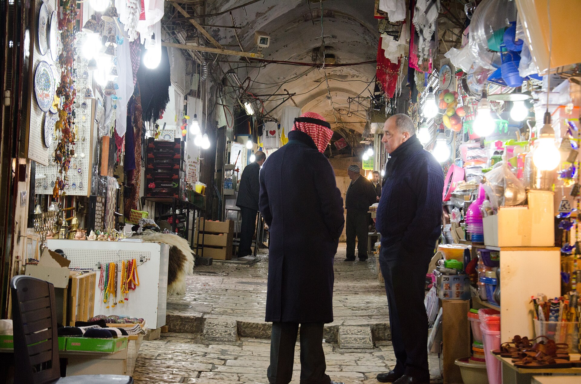 Two men in coats talk in a vaulted lane of Jerusalem's Old City souk, between brightly lit shops selling ceramic plates, prayer beads and housewares