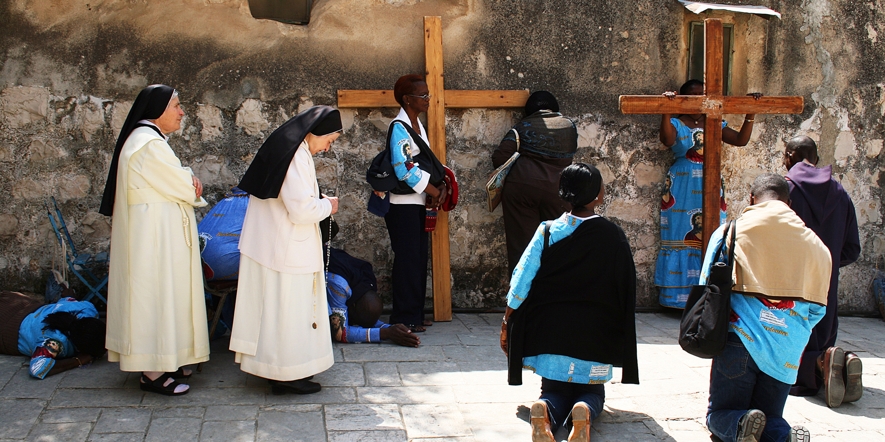 Two nuns in white habits standing in prayer as pilgrims in matching blue dresses kneel beside two large wooden crosses on the Via Dolorosa on Good Friday