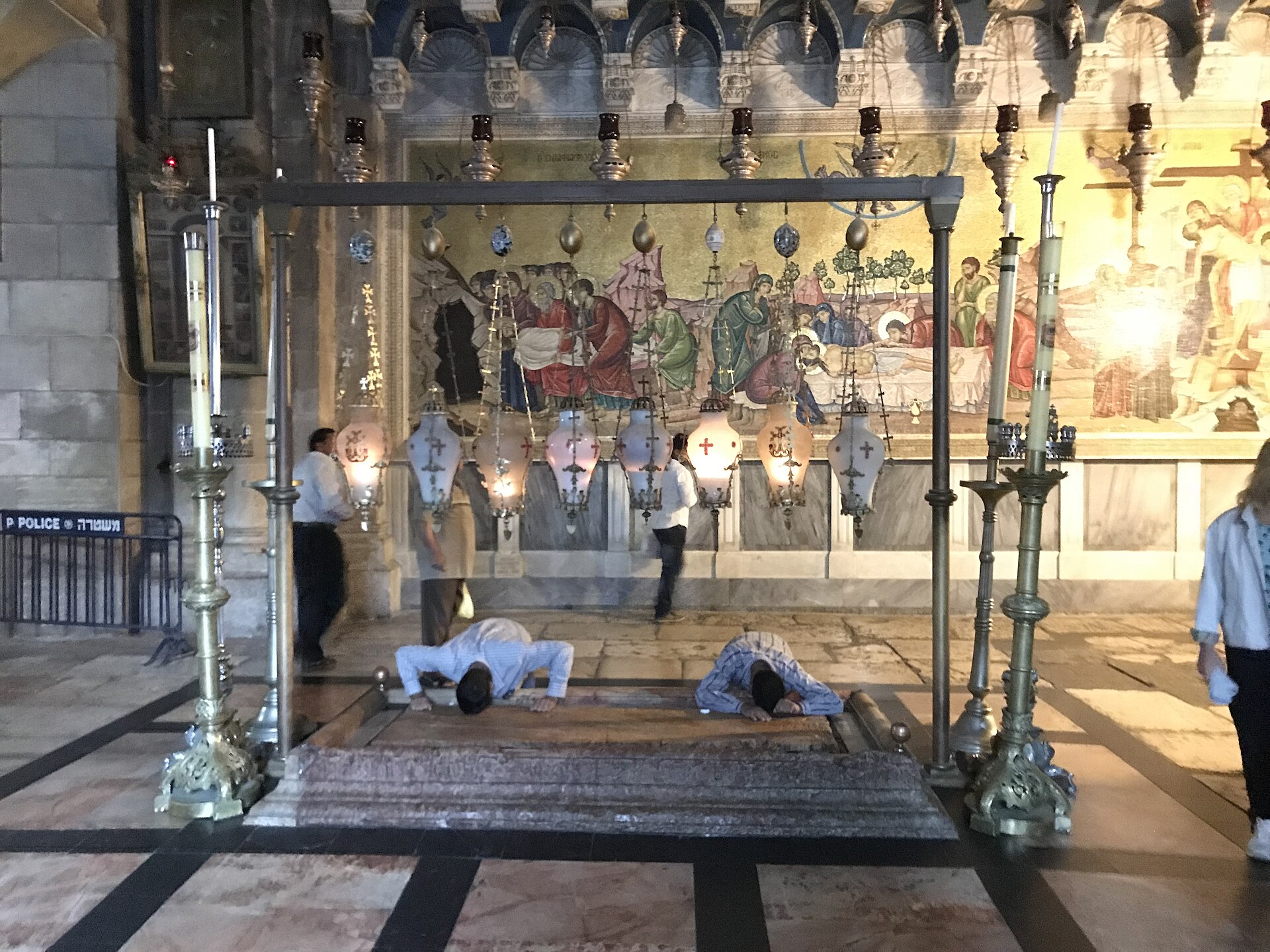 Two pilgrims bowing to touch the Stone of Anointing between tall candlesticks and hanging lamps, in front of a gold mosaic of Jesus's body being prepared for burial