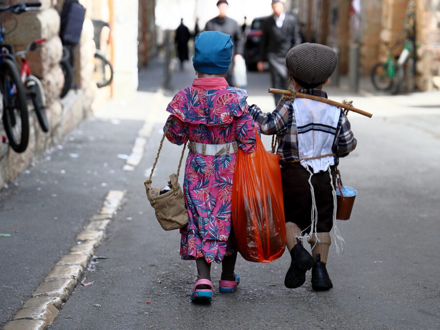 Two small children in Purim costumes, seen from behind, carry bags and a small pail down a confetti-strewn lane in Mea Shearim, Jerusalem