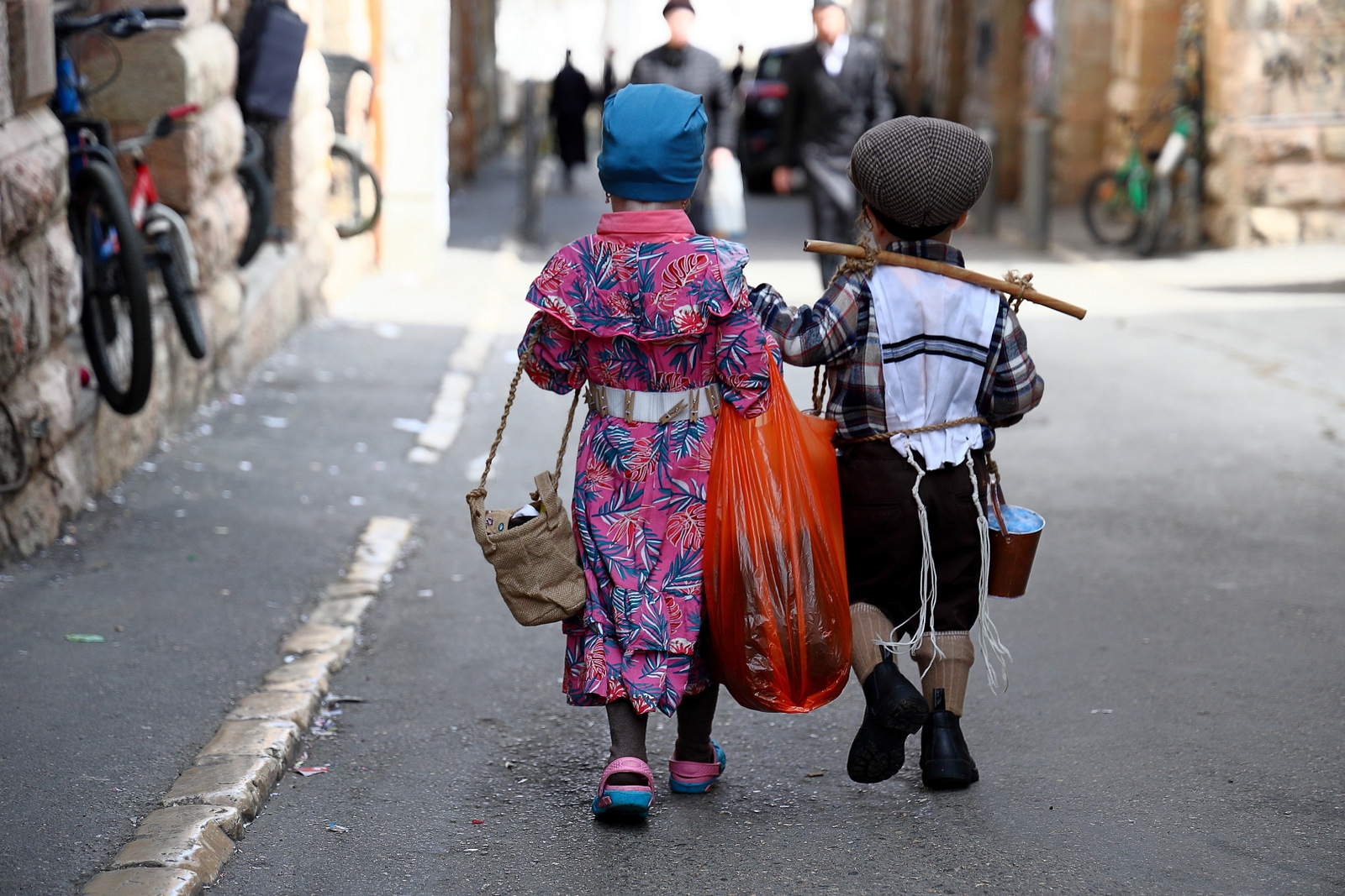 Two small children in Purim costumes, seen from behind, carry bags and a small pail down a confetti-strewn lane in Mea Shearim, Jerusalem