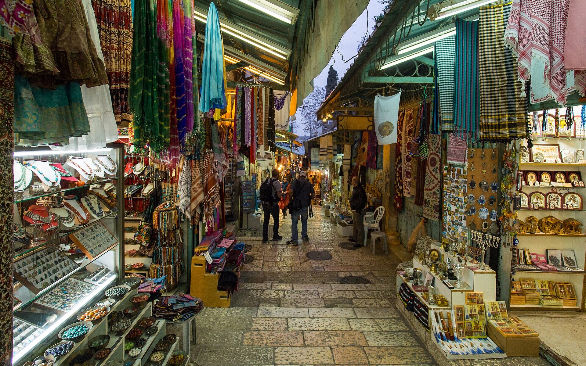 Two visitors with backpacks walk down David Street in Jerusalem's Old City, between souvenir stalls hung with scarves, jewellery, icons and menorahs