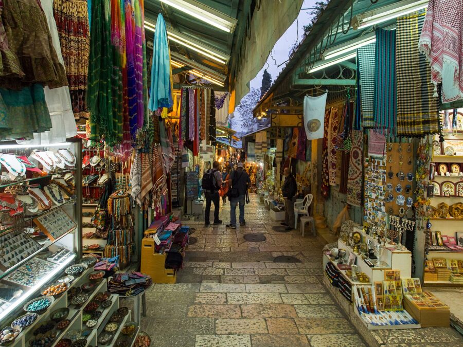 Two visitors with backpacks walk down David Street in Jerusalem's Old City, between souvenir stalls hung with scarves, jewellery, icons and menorahs