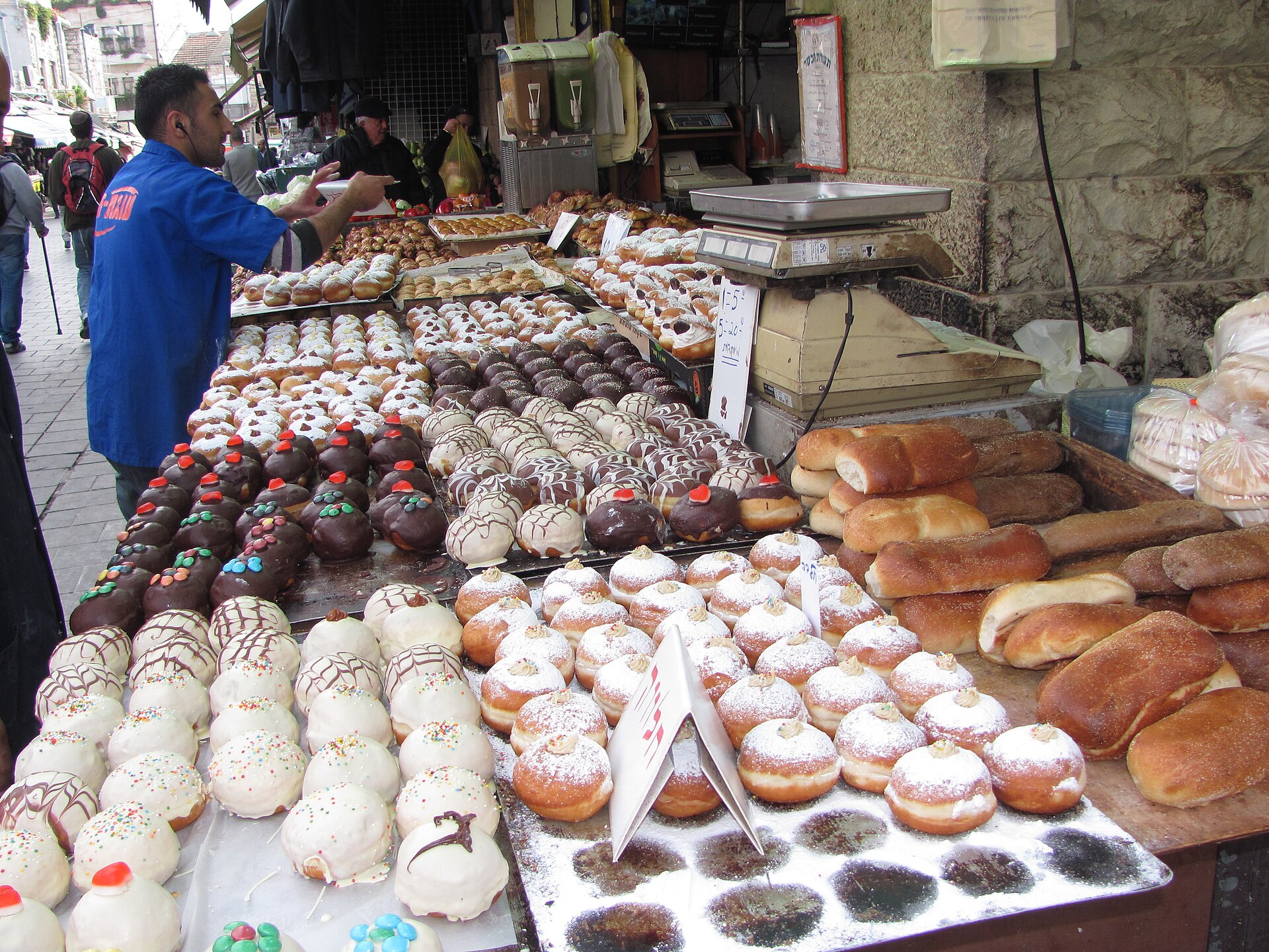 Vendor behind a Mahane Yehuda market stall piled with trays of sufganiyot glazed in chocolate or white icing with sprinkles, or dusted with sugar