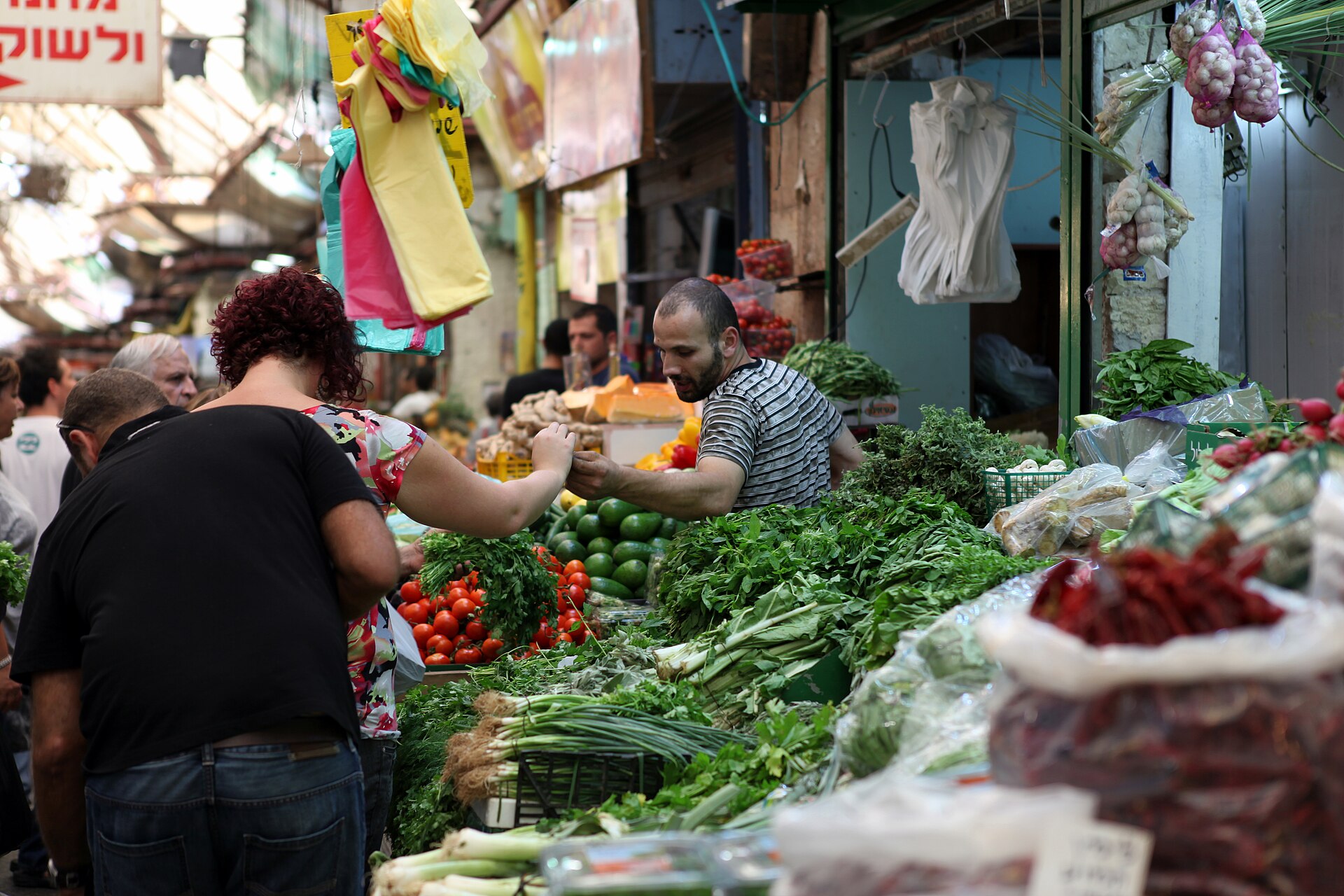 A vendor hands a customer a bunch of fresh herbs at a Mahane Yehuda Market stall piled with greens, spring onions, tomatoes and cucumbers, with garlic hanging overhead