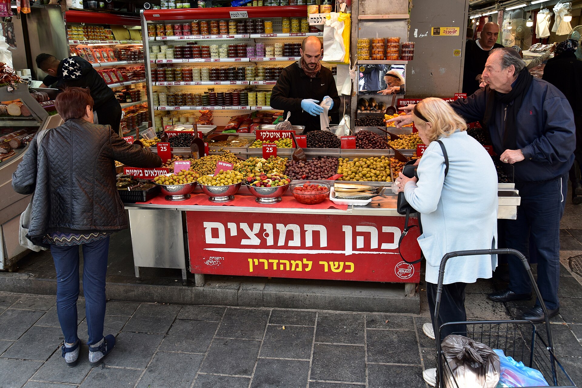 A vendor serves customers at the Cohen pickles stall in Mahane Yehuda Market, with trays of green and black olives, stuffed vegetables and a red Hebrew sign
