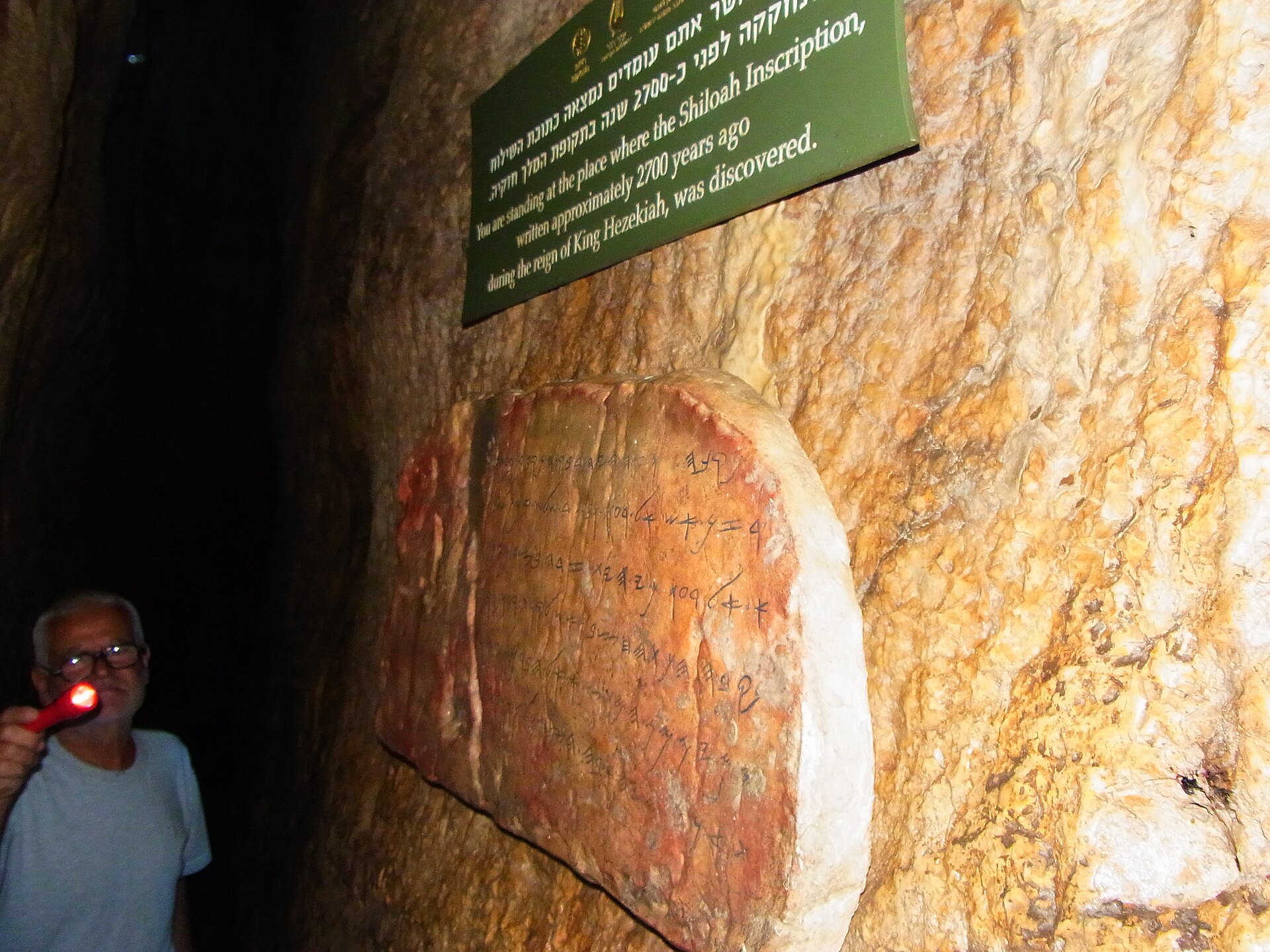 Visitor with a torch beside a replica of the Siloam Inscription on the rock wall of Hezekiah's Tunnel, below a Hebrew and English sign