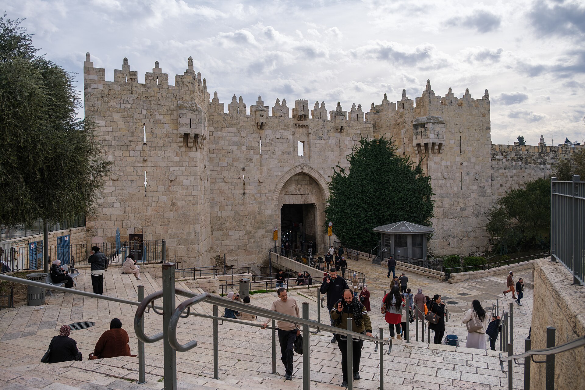 Visitors climbing the broad stone steps of the plaza in front of Damascus Gate, whose crenellated towers and pointed-arch entrance rise under a cloudy sky