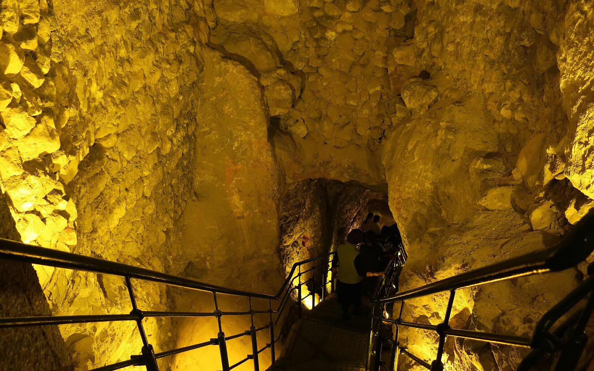 Visitors descending a railed metal staircase through a narrow, golden-lit rock-cut passage leading down into the Warren's Shaft water system
