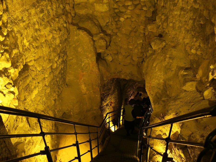 Visitors descending a railed metal staircase through a narrow, golden-lit rock-cut passage leading down into the Warren's Shaft water system