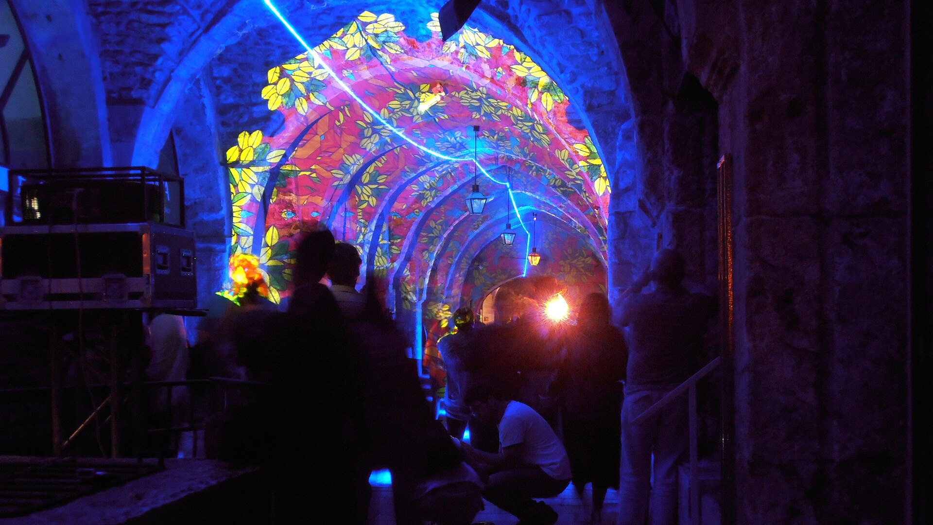 Visitors in silhouette walking under a vaulted stone arcade with hanging lanterns, its arches covered in projected leaf patterns in yellow, pink and blue