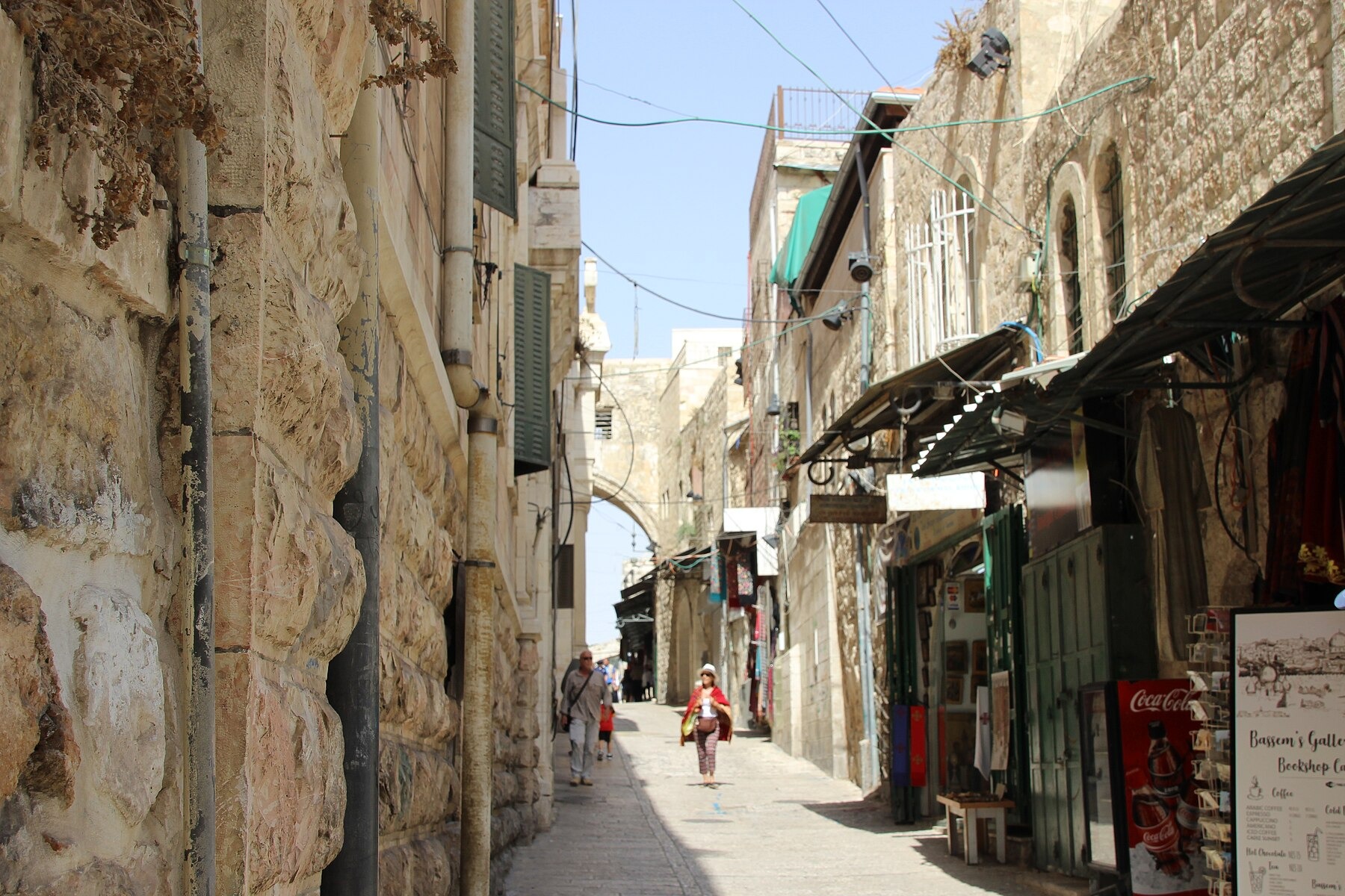 Visitors stroll along a narrow stretch of the Via Dolorosa in Jerusalem's Old City, past souvenir shops, with the Ecce Homo arch spanning the lane ahead