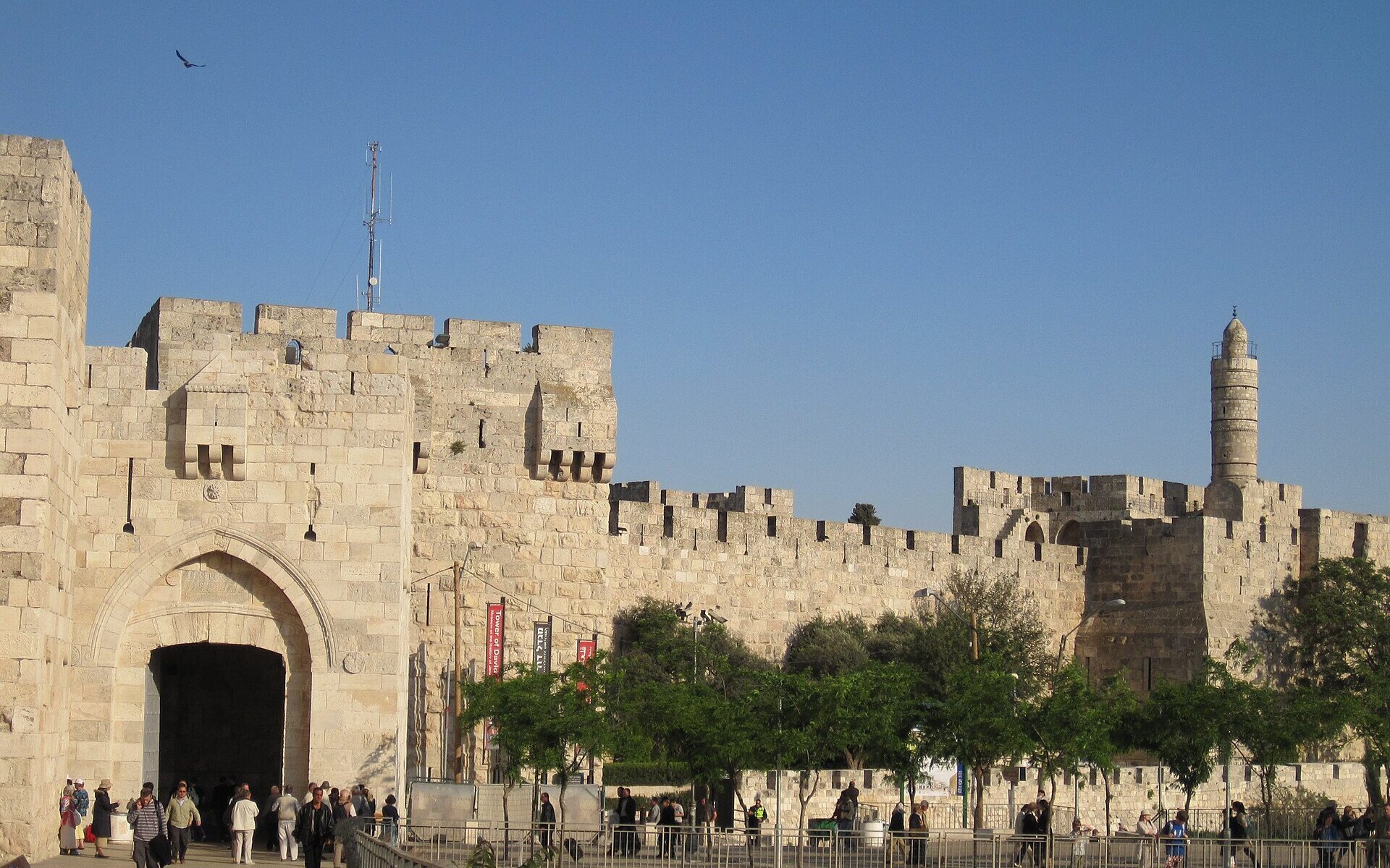 Visitors walking into Jaffa Gate's pointed-arch entrance, with the crenellated Old City wall stretching right to the Tower of David citadel and its slender minaret