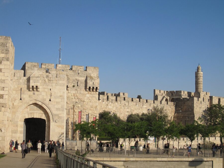 Visitors walking into Jaffa Gate's pointed-arch entrance, with the crenellated Old City wall stretching right to the Tower of David citadel and its slender minaret