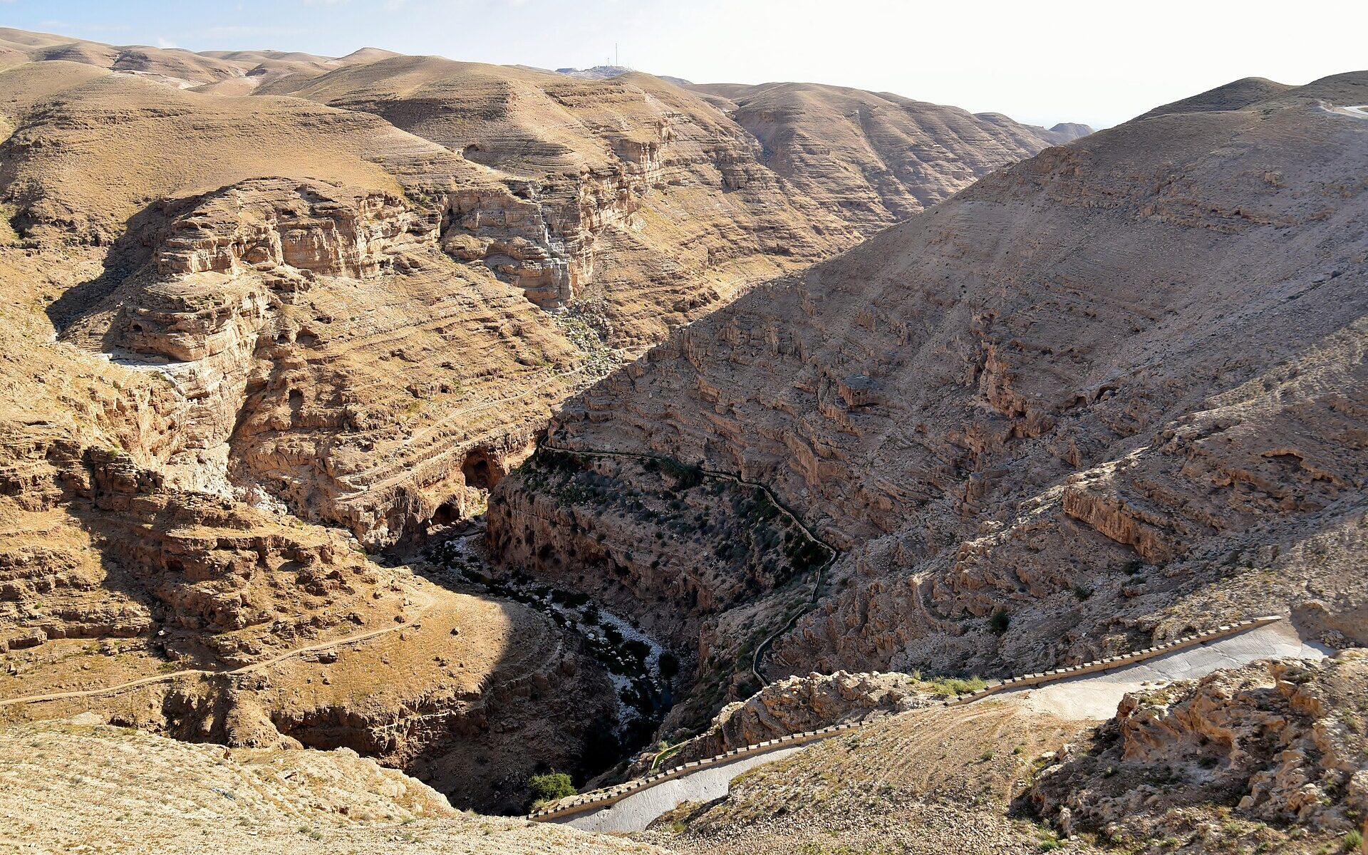 Wadi Qelt gorge in the Judean Desert seen from above, with layered limestone cliffs, a green streambed, the aqueduct channel along one slope and a paved path descending