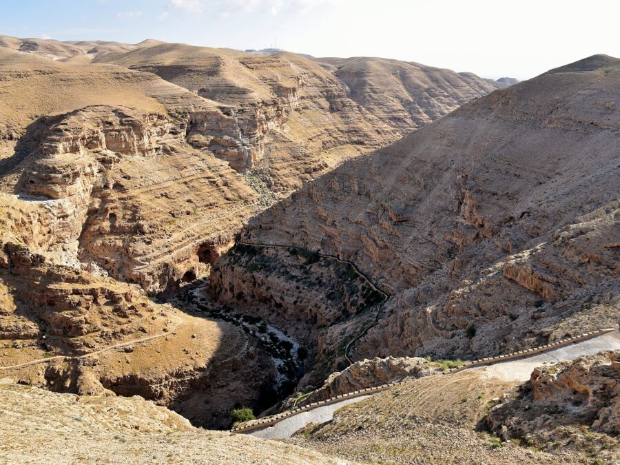 Wadi Qelt gorge in the Judean Desert seen from above, with layered limestone cliffs, a green streambed, the aqueduct channel along one slope and a paved path descending