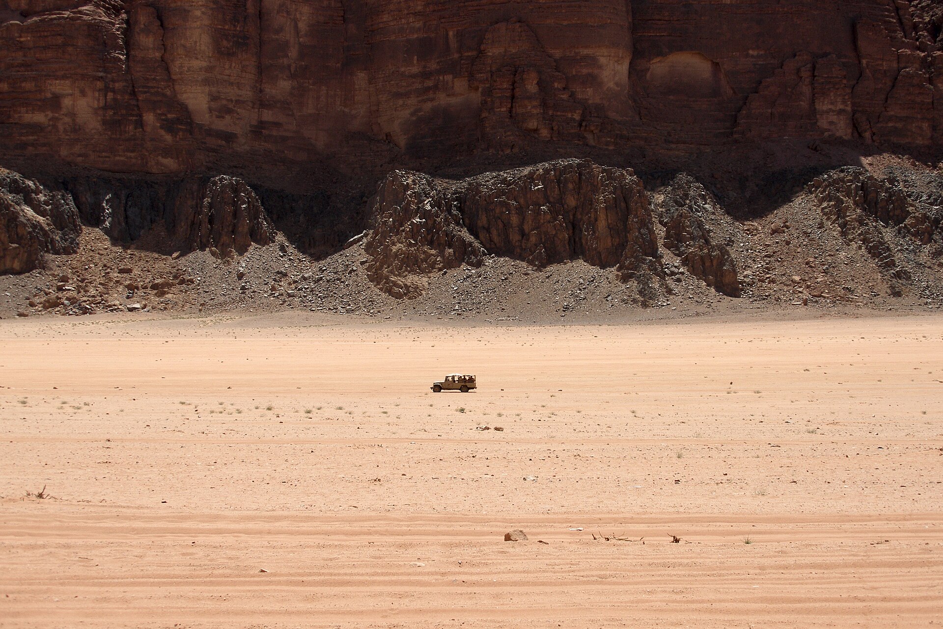 A lone four-wheel-drive vehicle crossing the flat pink sand of Wadi Rum in Jordan beneath towering dark sandstone cliffs