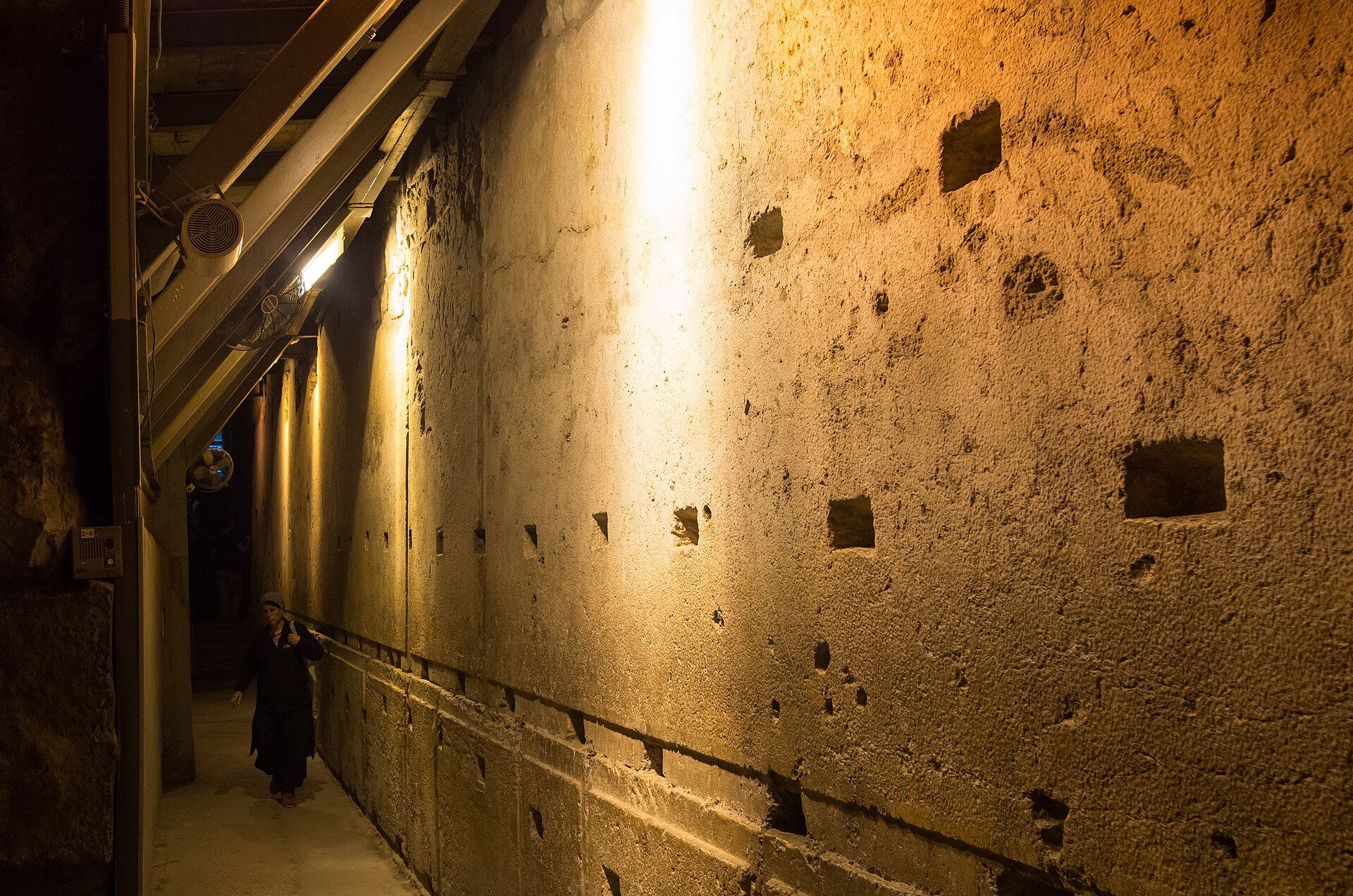 The Western Stone, a single enormous Herodian block pitted with square sockets, stretches along a lit passage of the Western Wall Tunnels past a walking visitor