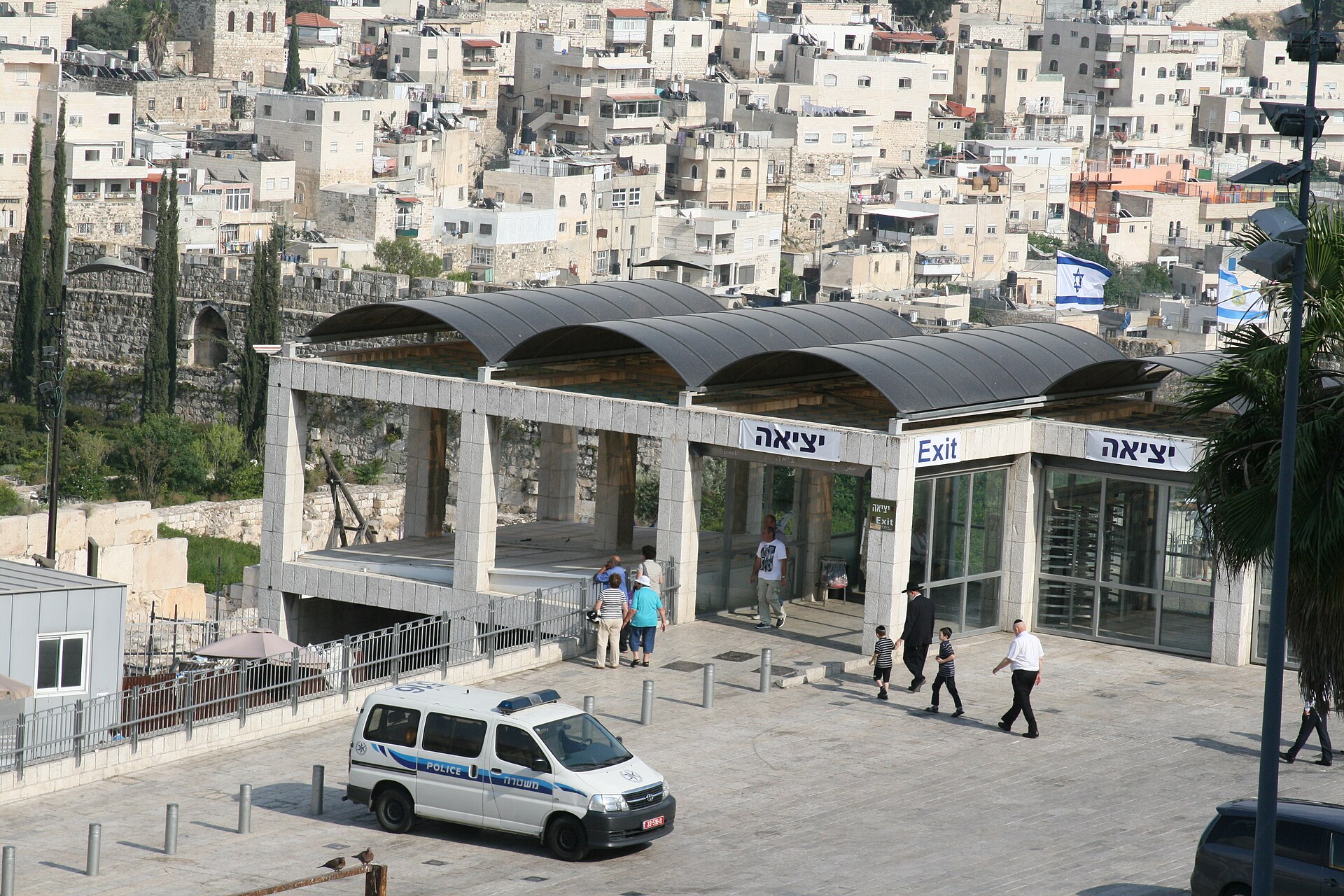 The Western Wall Gateway security entrance with turnstiles and bilingual Exit signs, a parked Israel Police van in front and visitors walking calmly across the plaza