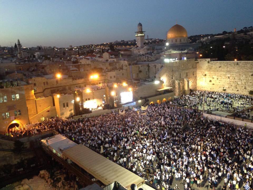 Aerial evening view of the Western Wall plaza packed with thousands of people carrying Israeli flags, with the Dome of the Rock lit behind