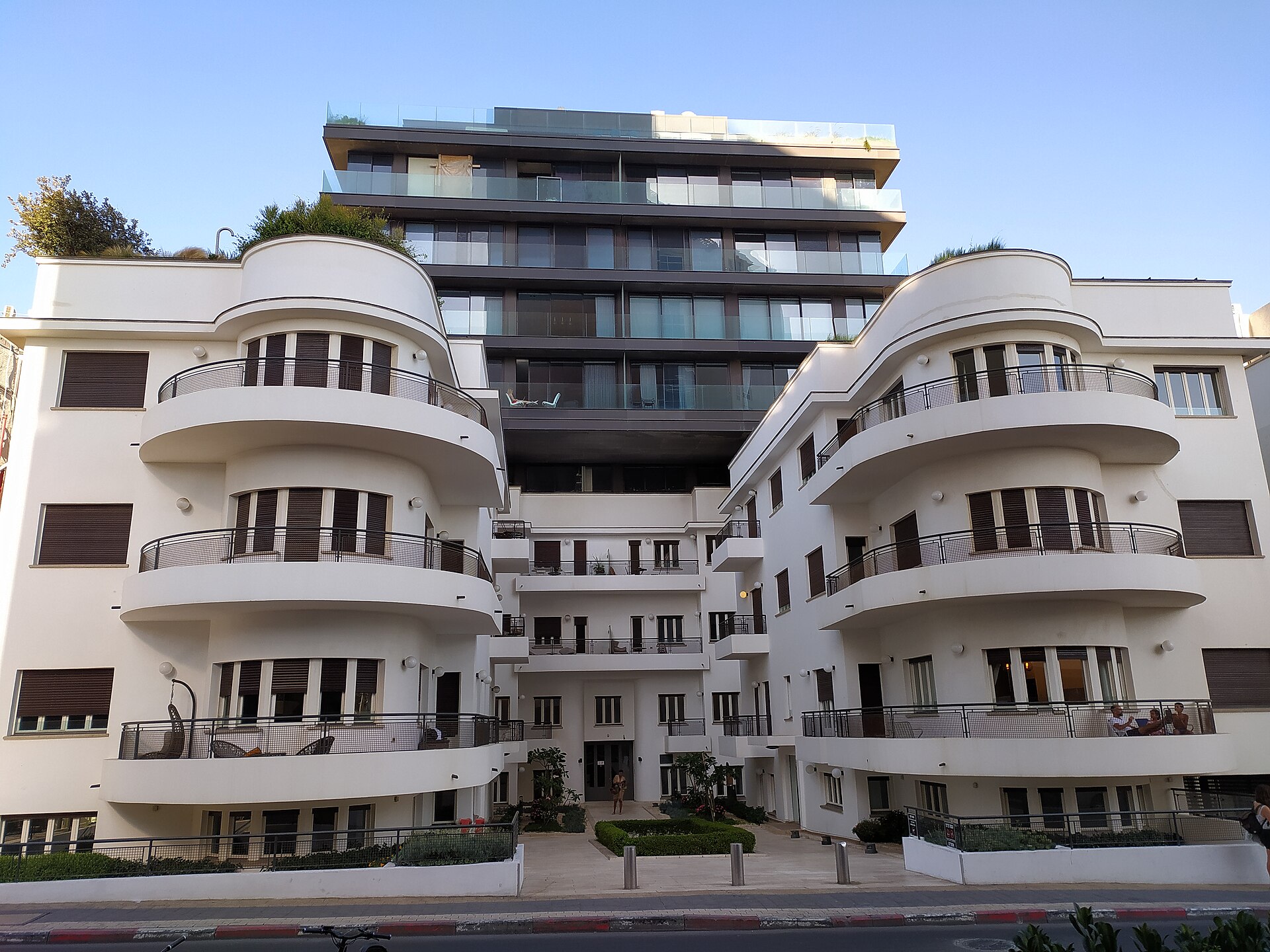 White 1930s International Style apartment building in Tel Aviv with two wings of stacked rounded balconies, with a modern glass tower rising behind it