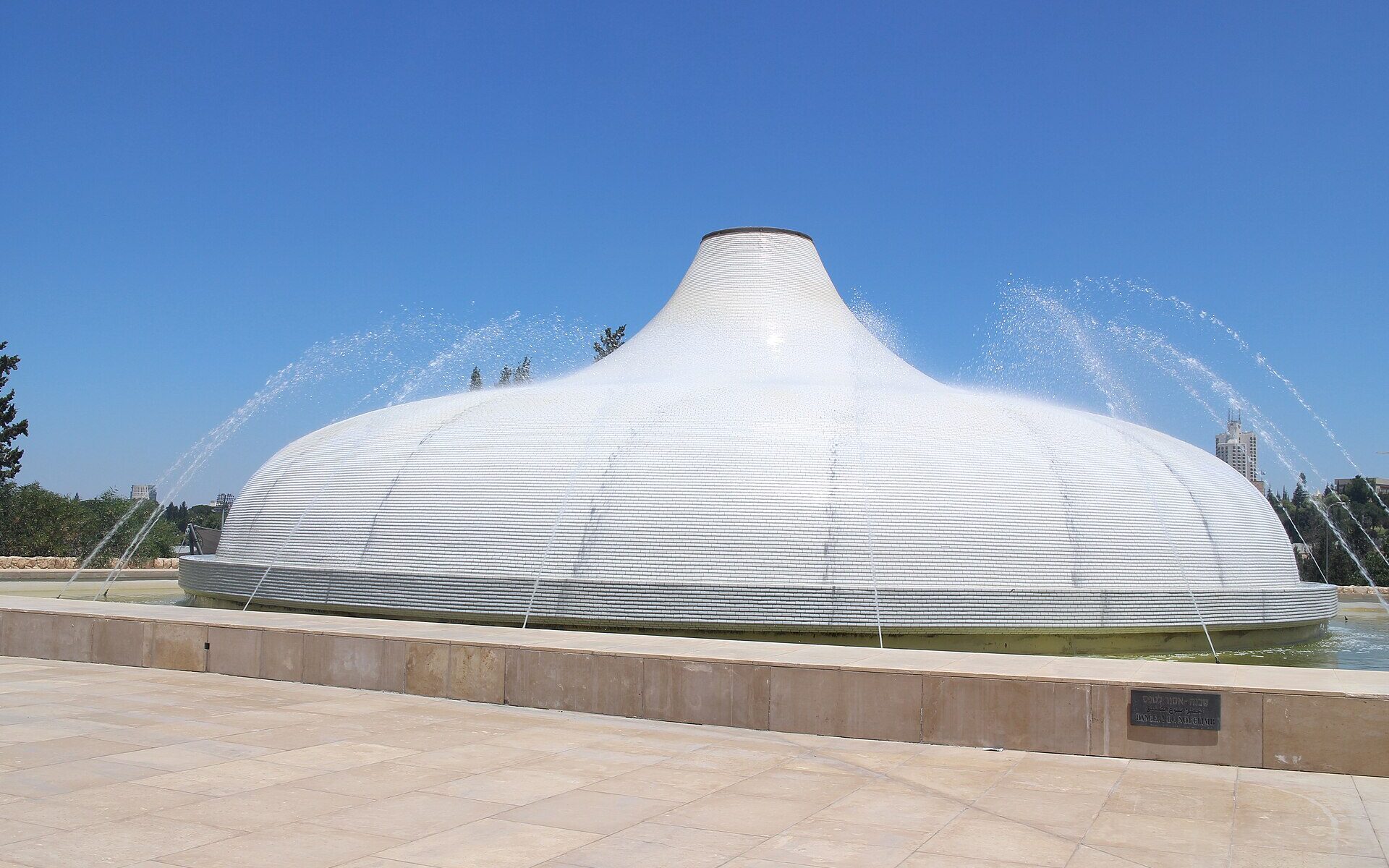 White tiled dome of the Shrine of the Book at the Israel Museum, shaped like a scroll-jar lid, with fountain jets arcing over it beyond a stone plaza