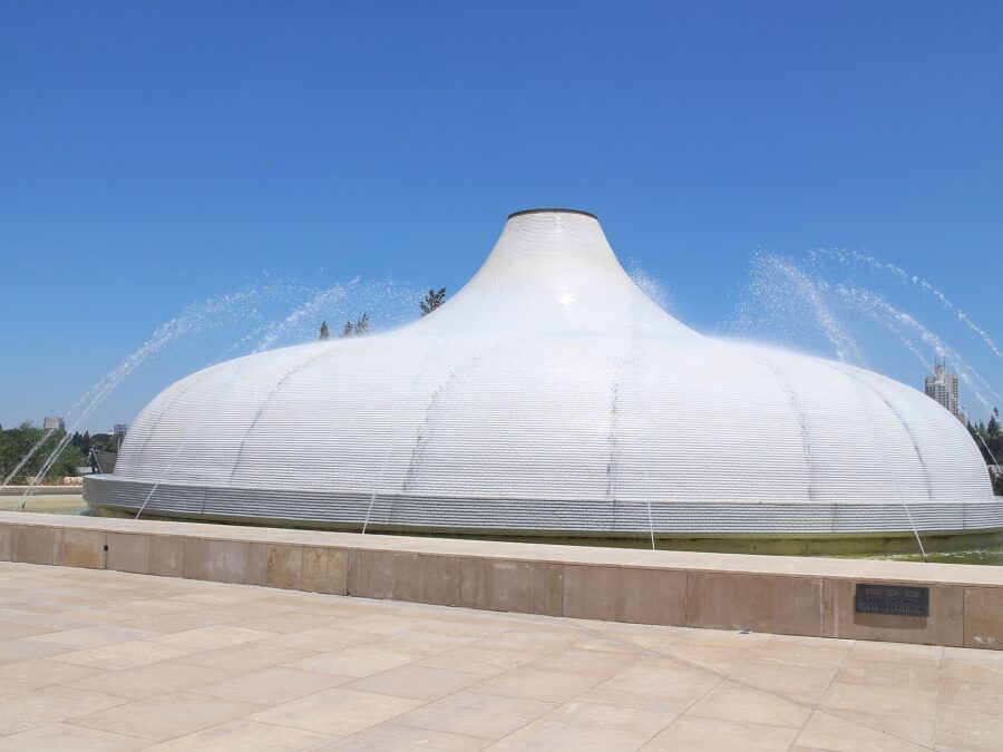 White tiled dome of the Shrine of the Book at the Israel Museum, shaped like a scroll-jar lid, with fountain jets arcing over it beyond a stone plaza