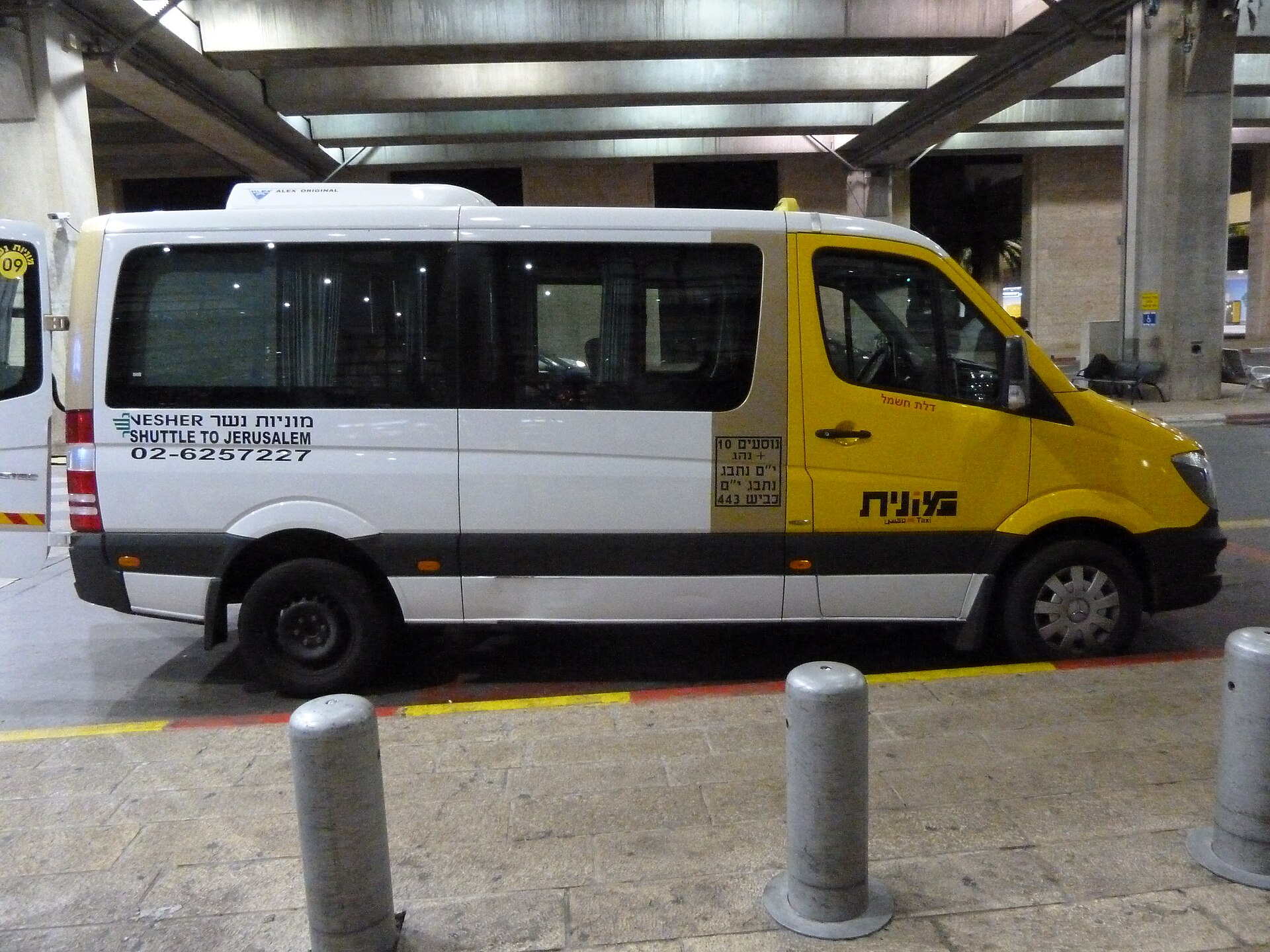 White and yellow Nesher shared-taxi minibus lettered Shuttle to Jerusalem in Hebrew and English, parked at the kerb outside Ben Gurion Airport at night