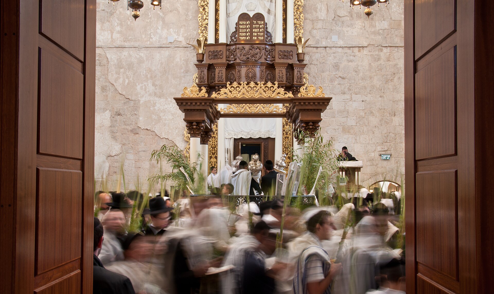 Worshippers in prayer shawls circle with palm-frond lulavim before the gilded Torah ark of the Hurva Synagogue during Hoshana Rabbah, seen through open doors