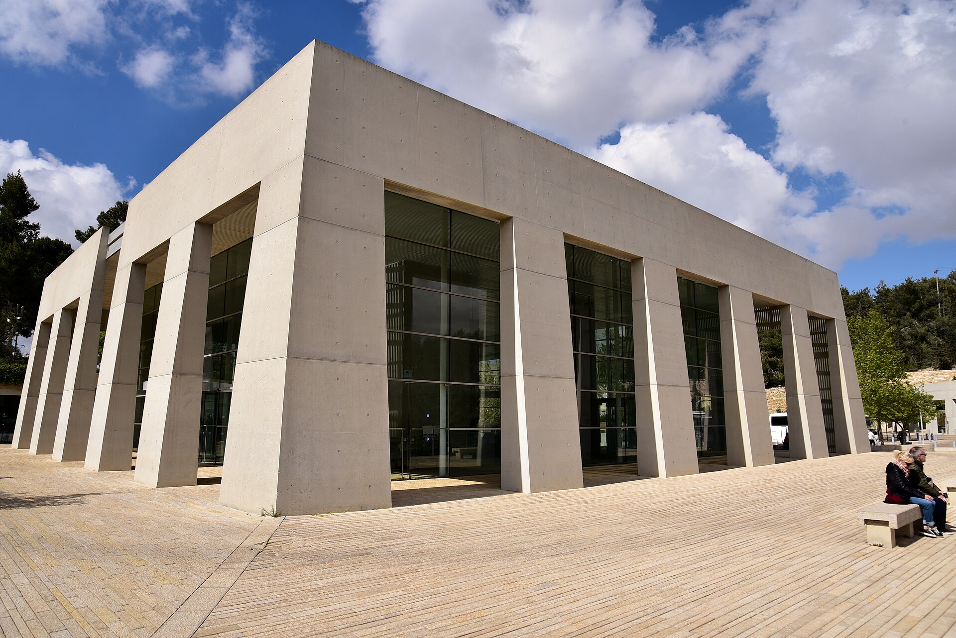 Yad Vashem's visitors' centre, a flat-roofed pavilion of tall concrete piers and glass walls standing on a stone-paved plaza, with two people resting on a bench