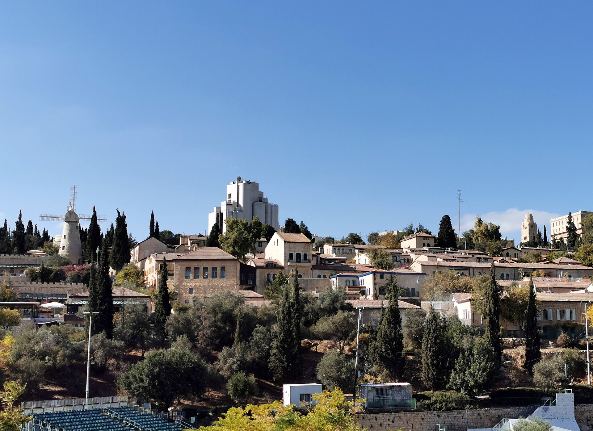 Yemin Moshe's terraced stone houses with tiled roofs among cypresses and olive trees on a Jerusalem hillside, with the Montefiore Windmill at left